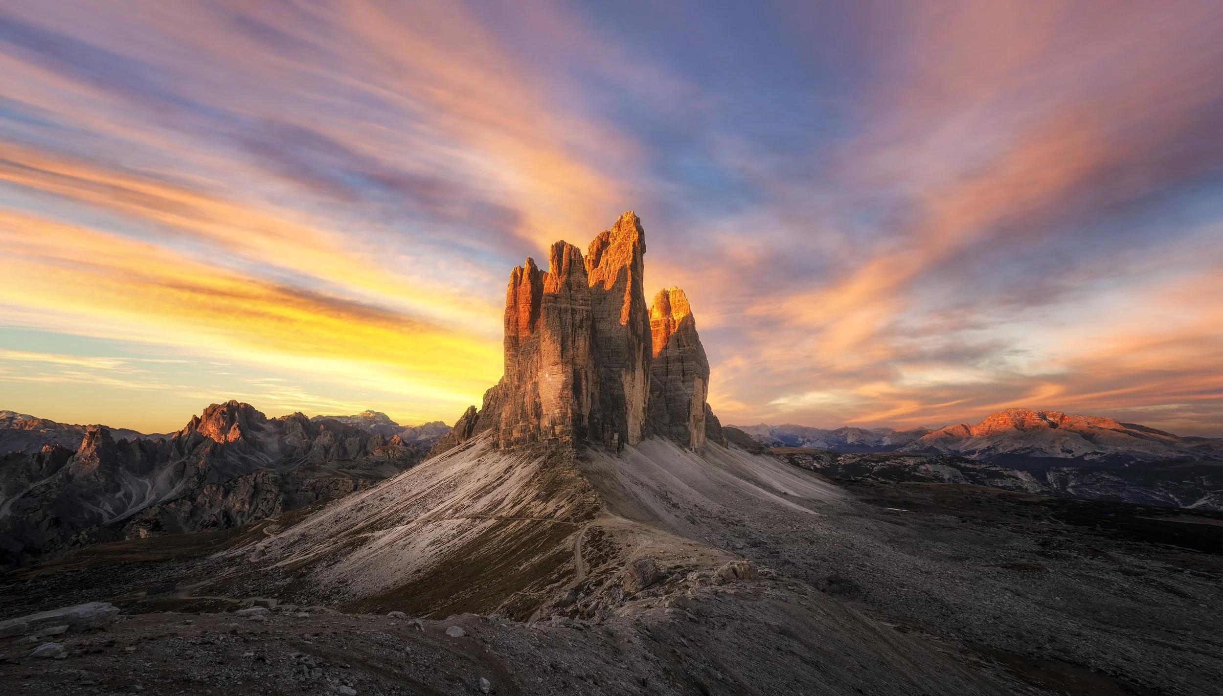 A mountain with a jagged peak during a colorful sunset, with a sky showing pink, orange, and purple clouds and a rugged landscape in the foreground.