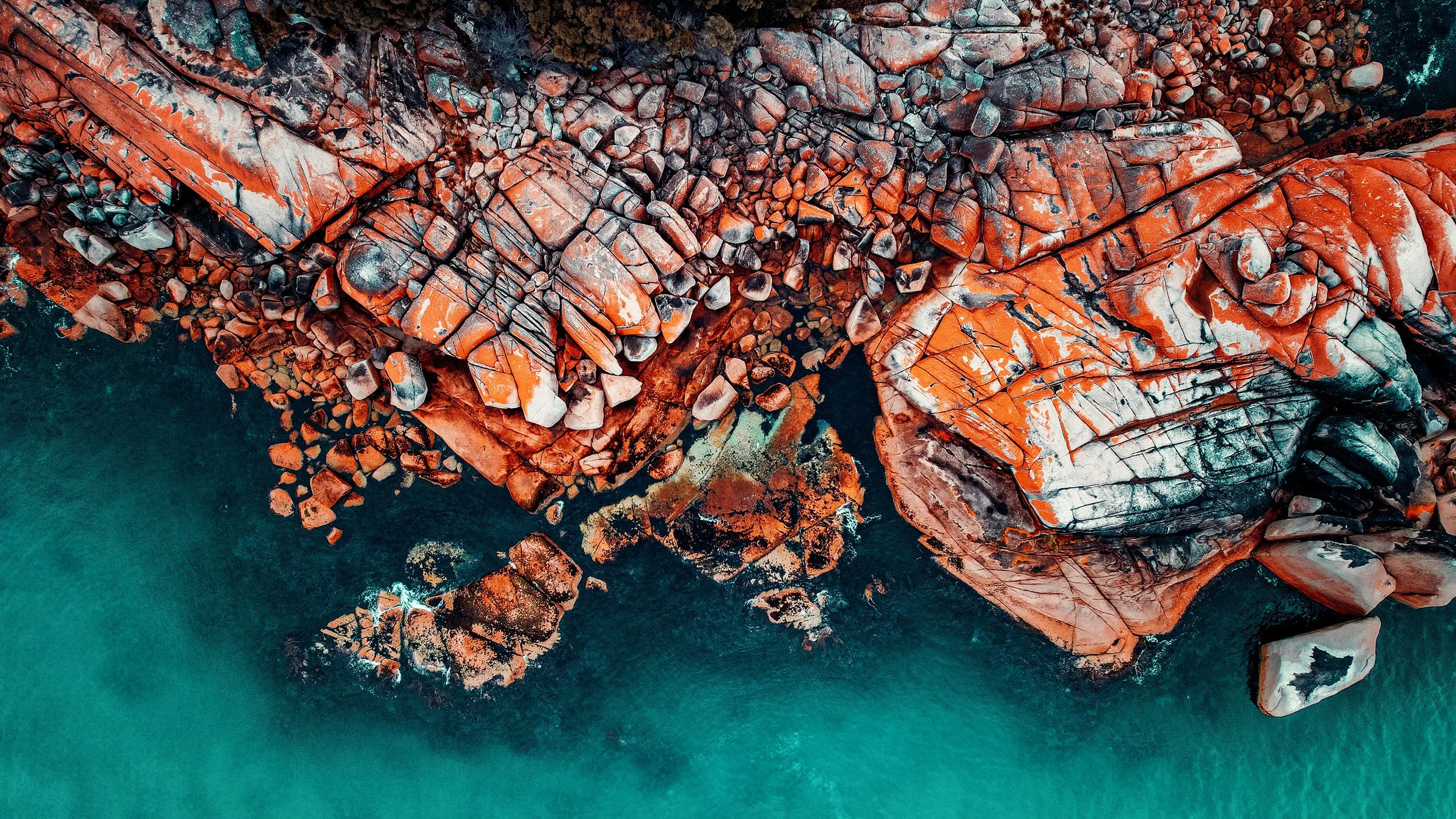 Aerial view of red and orange granite rocks along a turquoise shoreline.