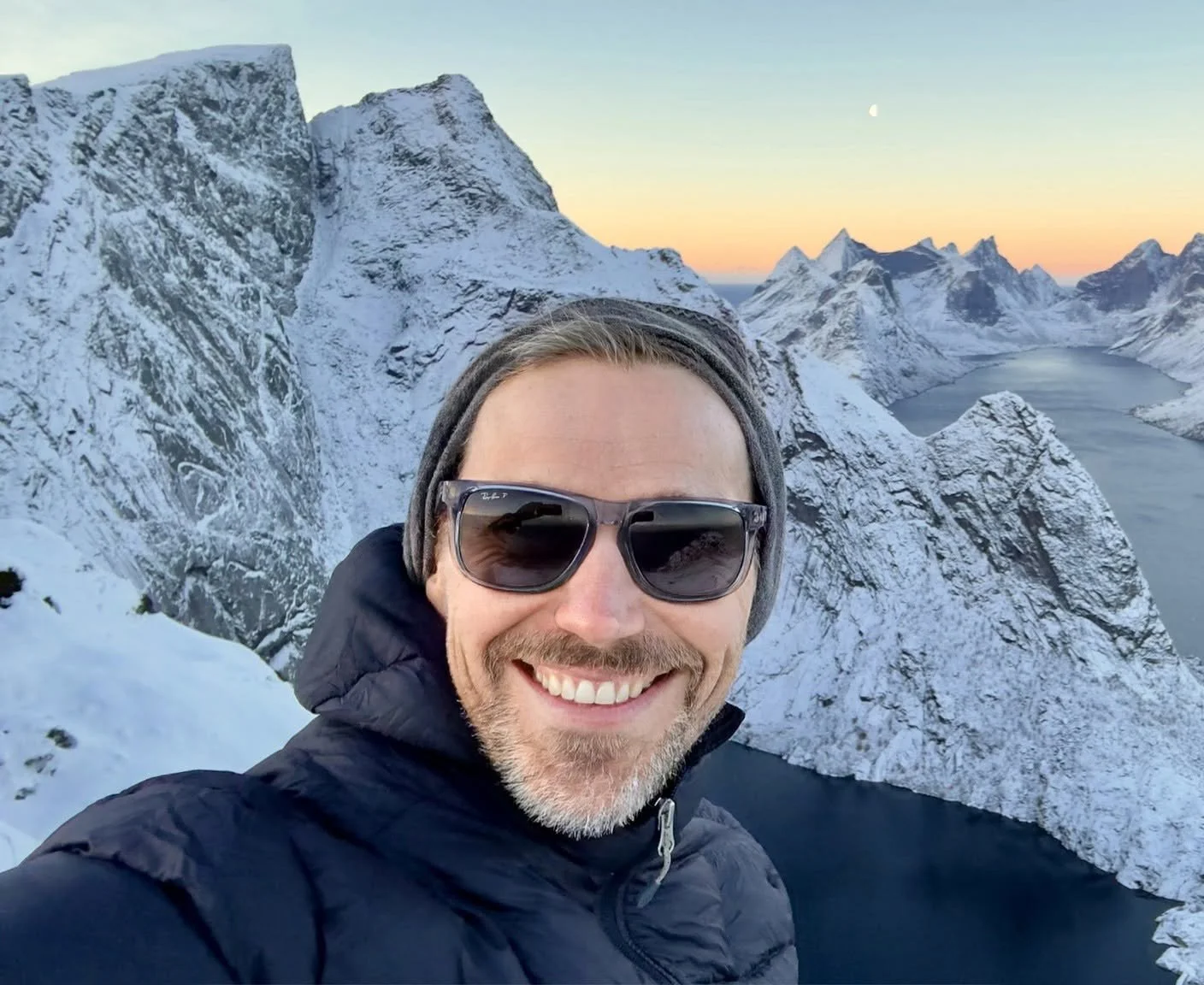 A smiling man wearing sunglasses, a beanie, and a dark jacket taking a selfie in a snowy mountainous landscape during sunset with a lake below.