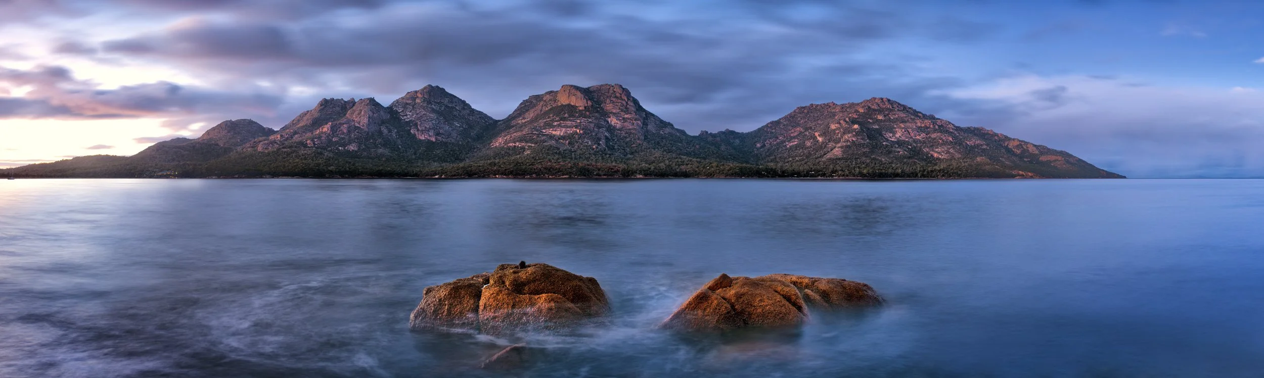A mountainous island view with rocky shores, calm water, and a cloudy sky at dusk.
