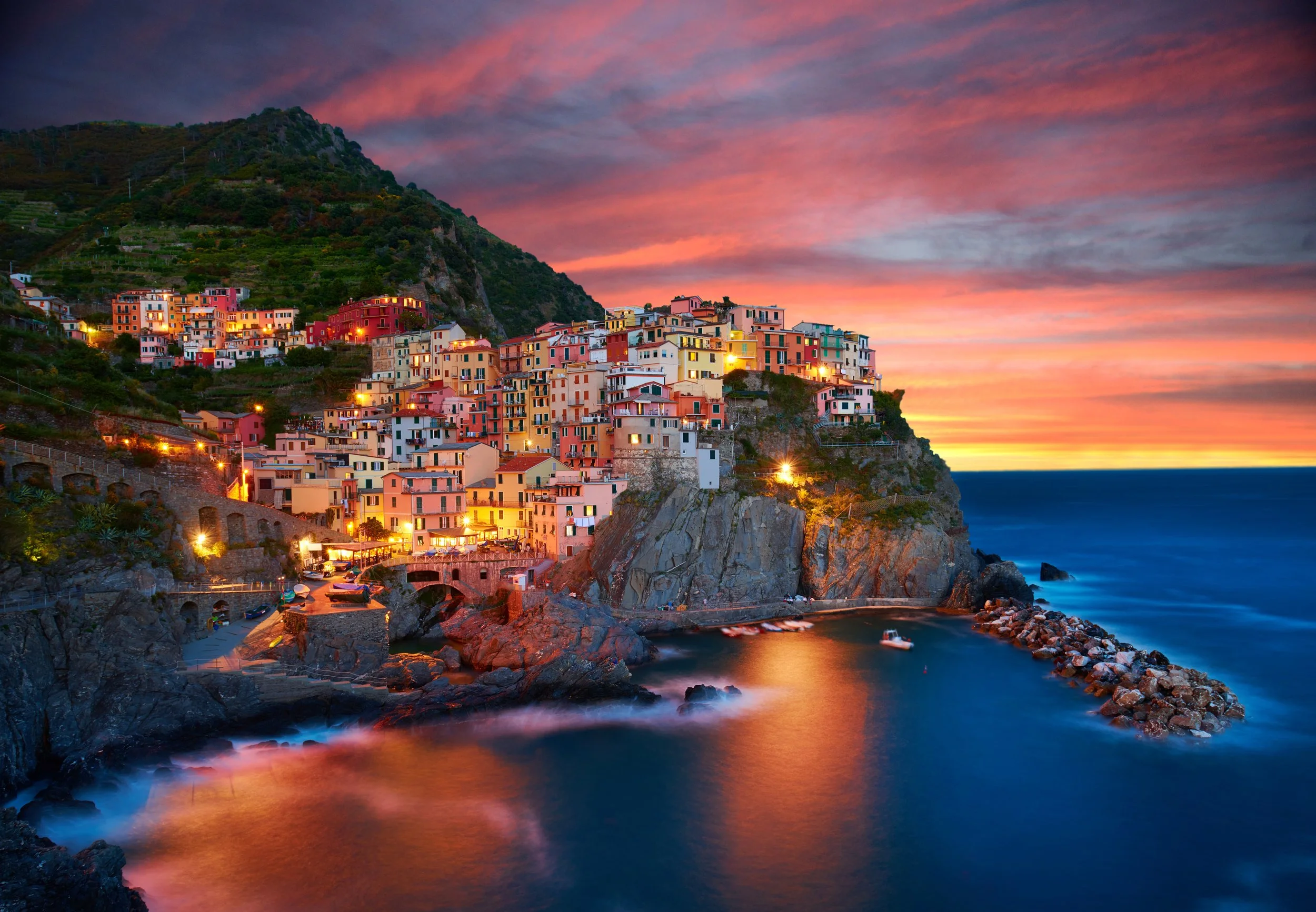 A colorful seaside village perched on a cliff at sunset, with boats in the water and the sky painted in pink, orange, and purple hues.