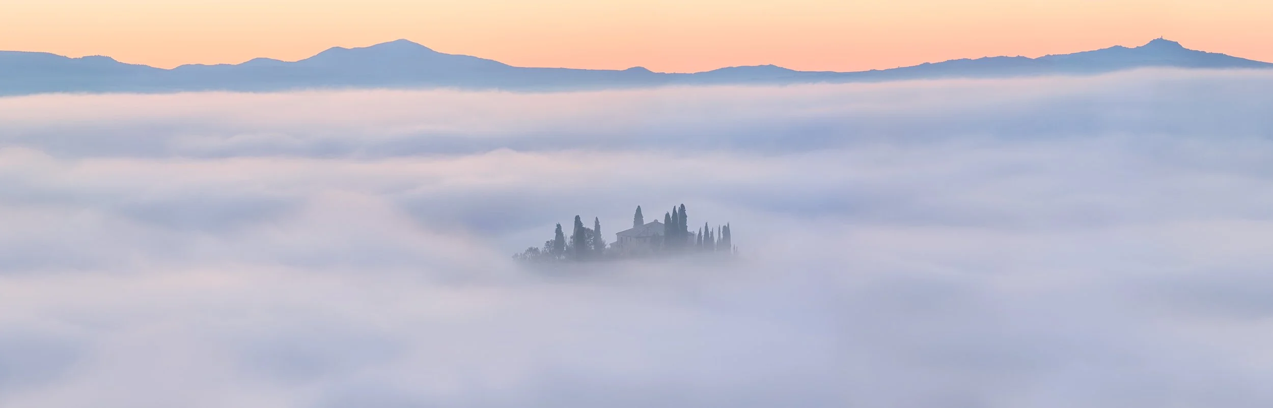 A mountain landscape with a building surrounded by cypress trees, partially obscured by thick fog, at sunrise or sunset.