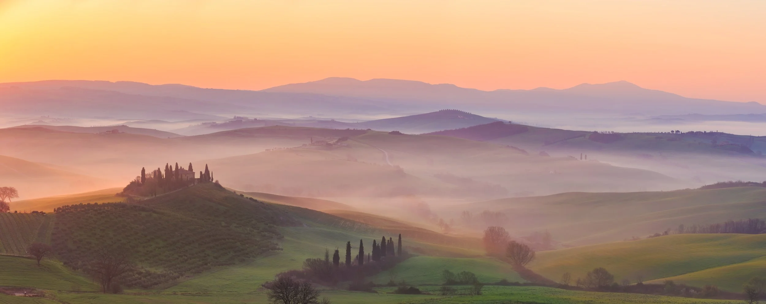 Scenic view of rolling hills at sunrise with layers of mist and trees, typical of Tuscany in Italy.