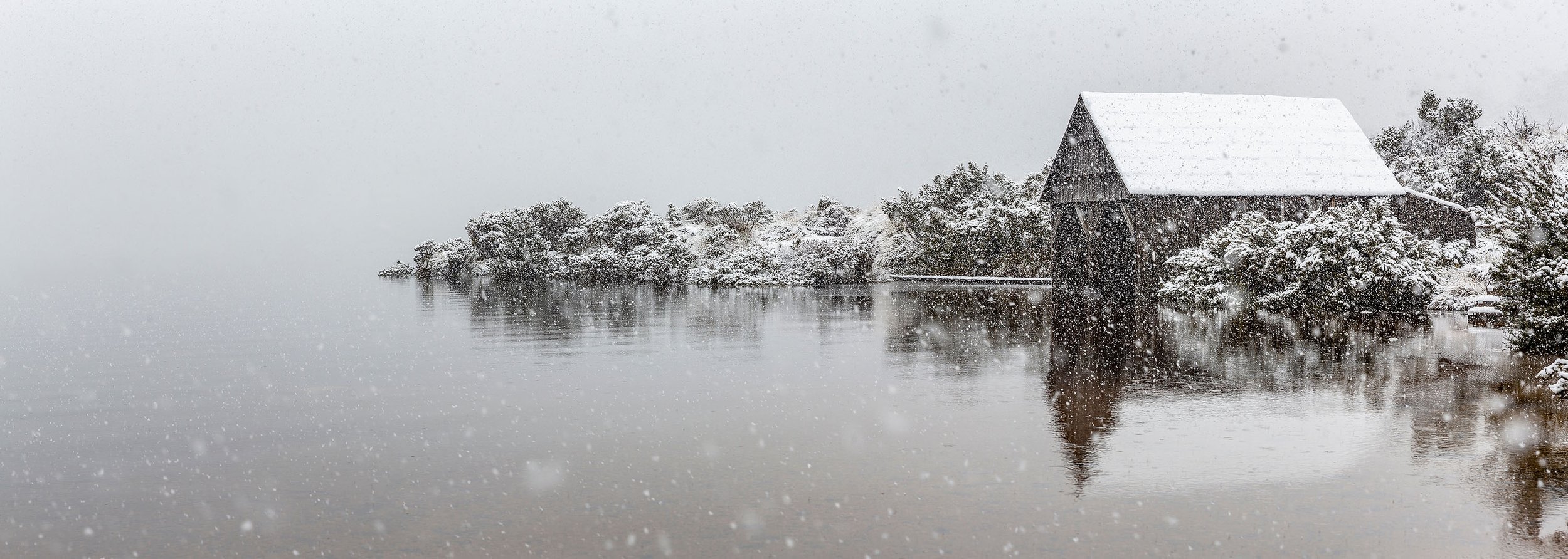 A snowy landscape featuring a barn by a lake, with snow-covered trees and falling snowflakes.