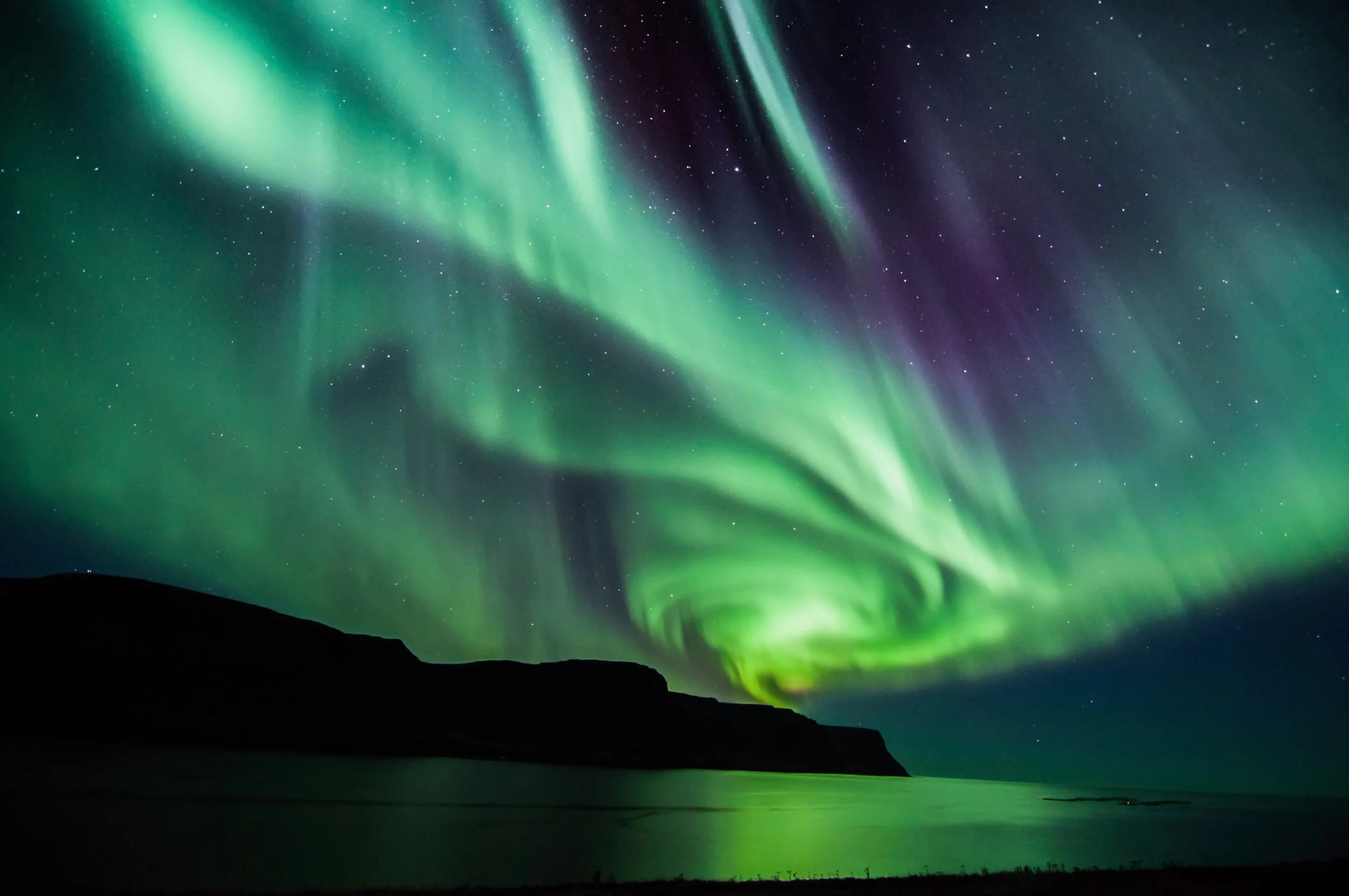 Northern lights display with green and purple streaks in the night sky over a calm body of water and silhouetted land.