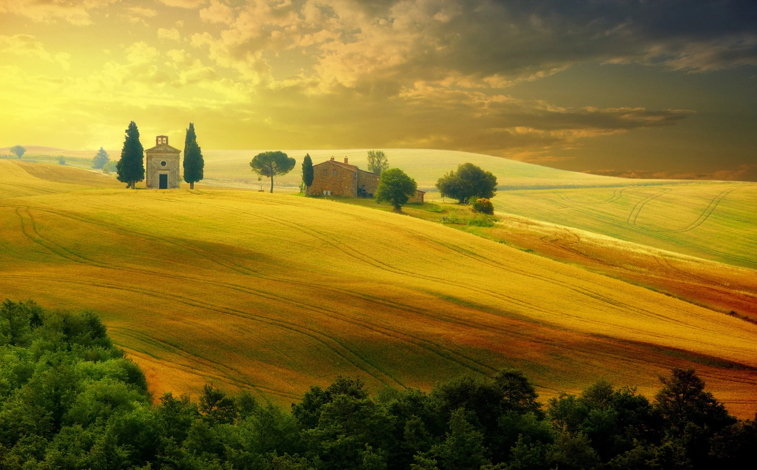 Sunset over a rolling countryside with a small chapel, trees, and fields.
