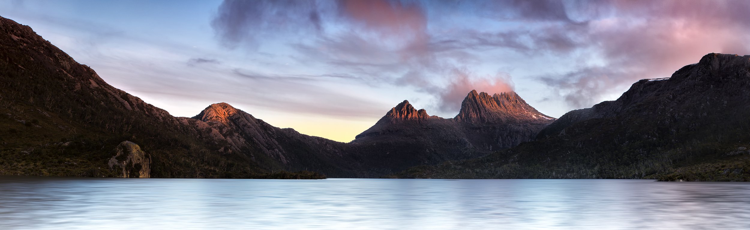 A serene mountain landscape with a calm lake in the foreground, surrounded by rugged peaks under a colorful, partly cloudy sky during sunset.