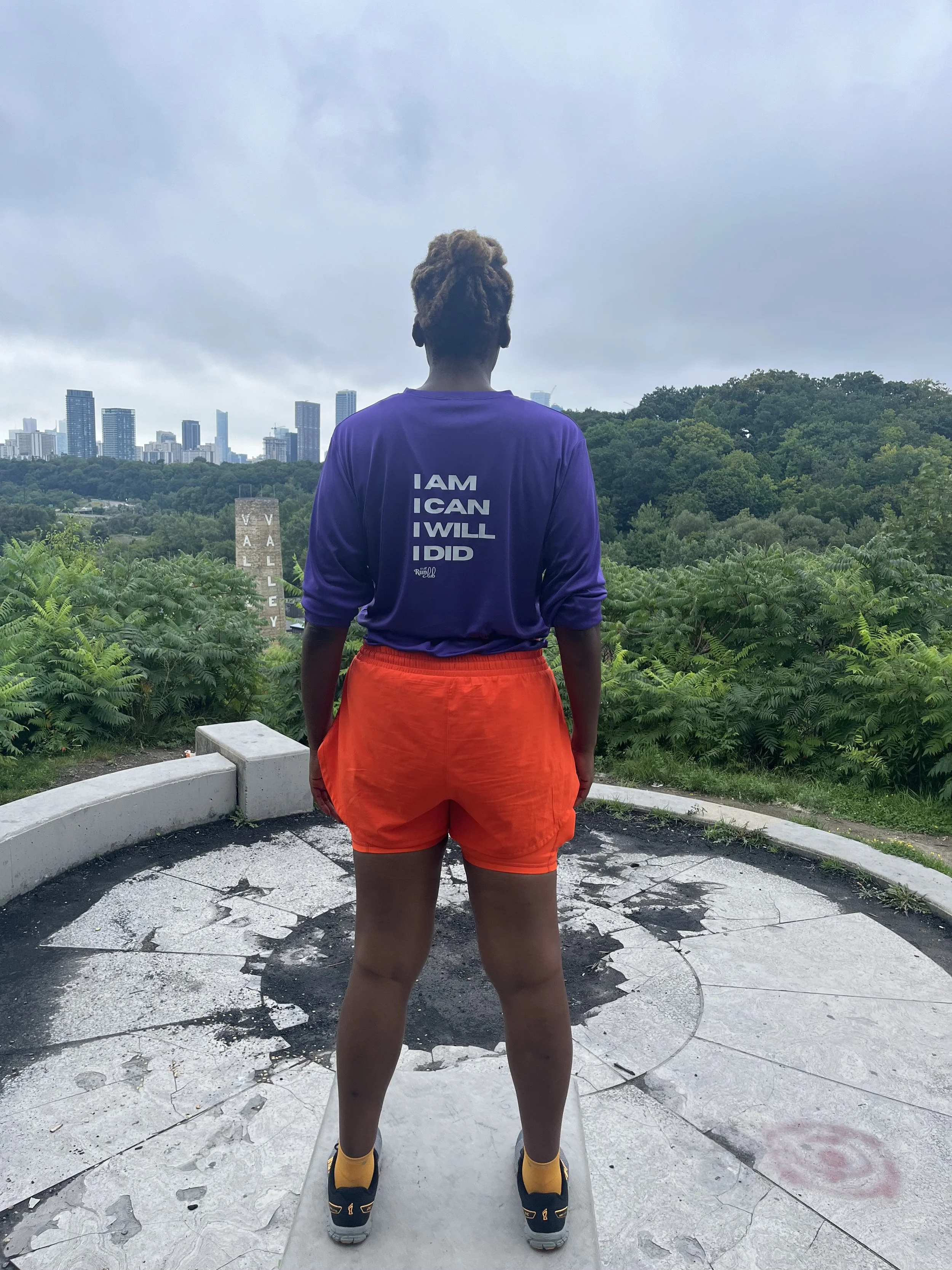 A person standing on an outdoor platform facing a cityscape, wearing a purple shirt with motivational words and orange shorts.