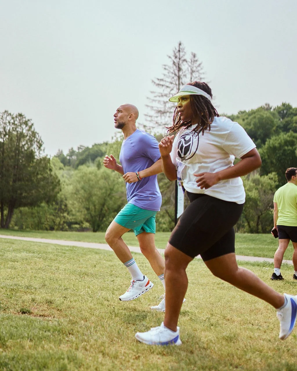 Two people running outdoors on a grassy field, surrounded by trees and greenery.