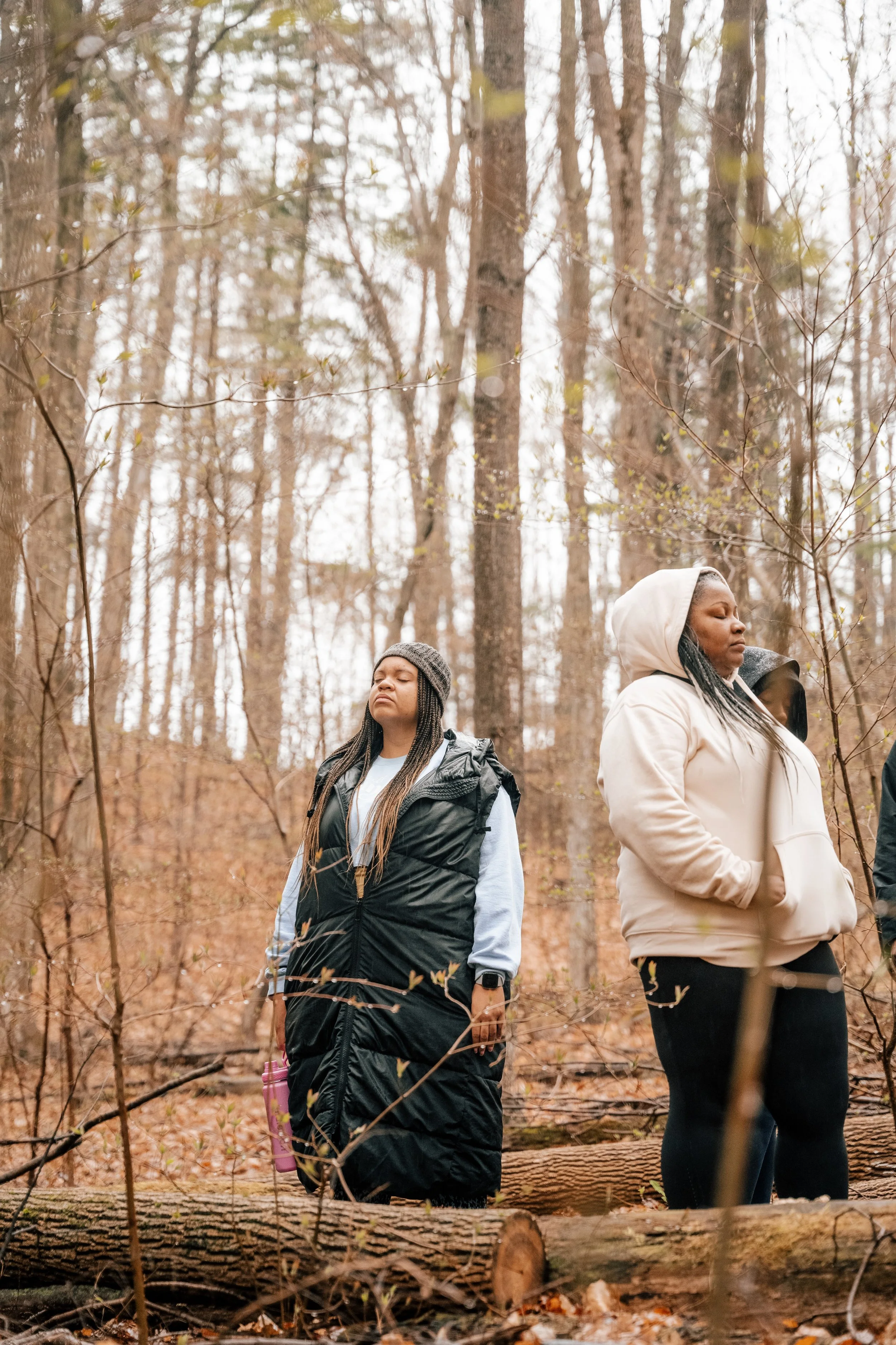 Two women in warm clothing stand in a forest with bare trees on a cloudy day, appearing to meditate or reflect.
