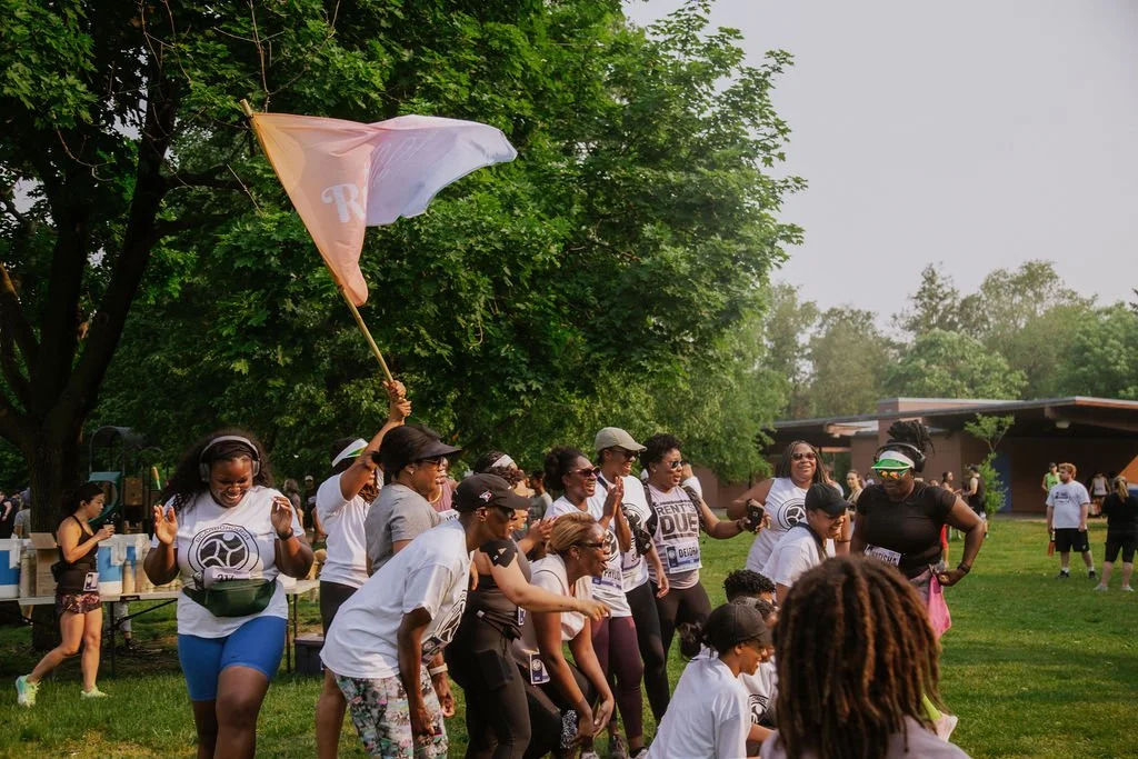 A group of women and girls gathered outdoors on a grassy area, participating in a team event or celebration, with some clapping and smiling, and a woman holding a flag.