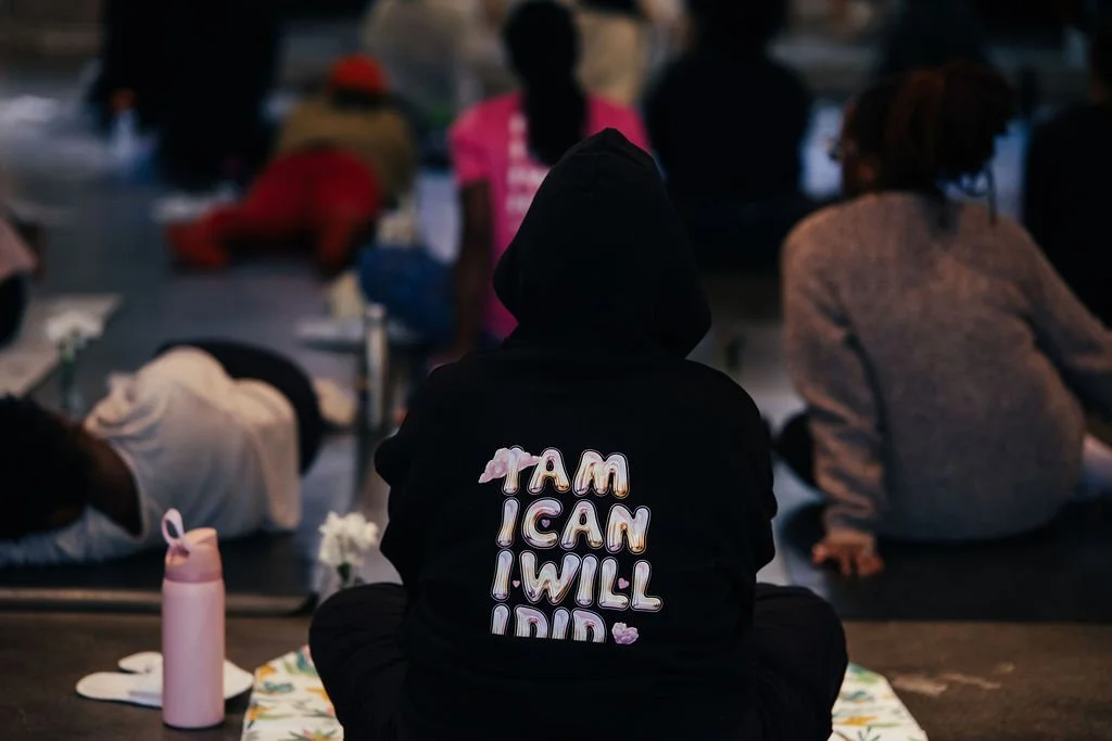 Person wearing a black hoodie with the words 'I AM I CAN I WILL' on the back, sitting with their back to the camera at a gathering or protest, surrounded by other people sitting or lying down.