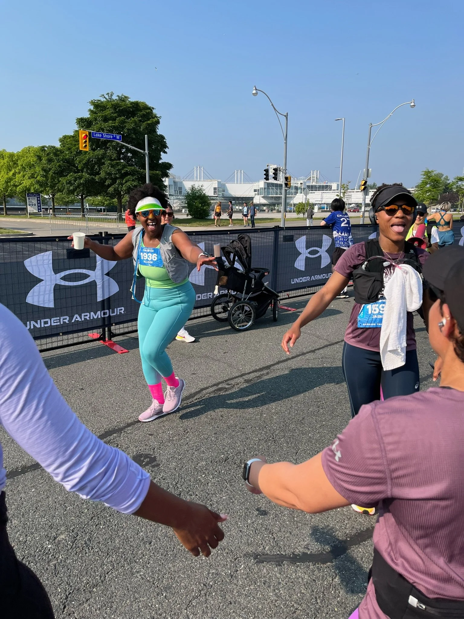 Two women participating in a running event, smiling and celebrating as they reach the finish line, with spectators and a barrier with Under Armour logos in the background, under a clear blue sky.