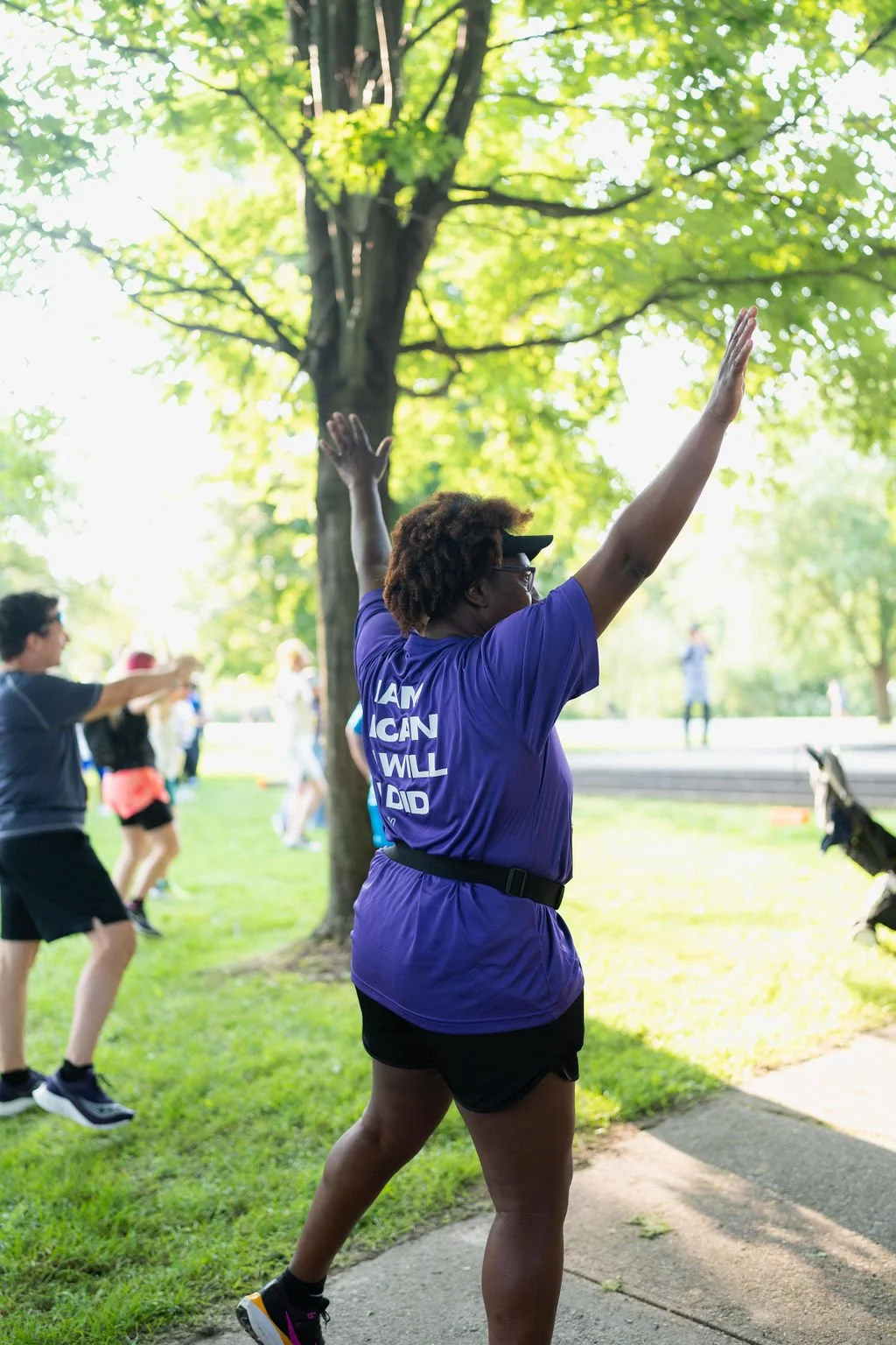 A group of people participating in a fitness class outdoors in a park, with one woman in the foreground stretching her arms up, wearing a purple shirt and shorts, and others behind her also exercising.