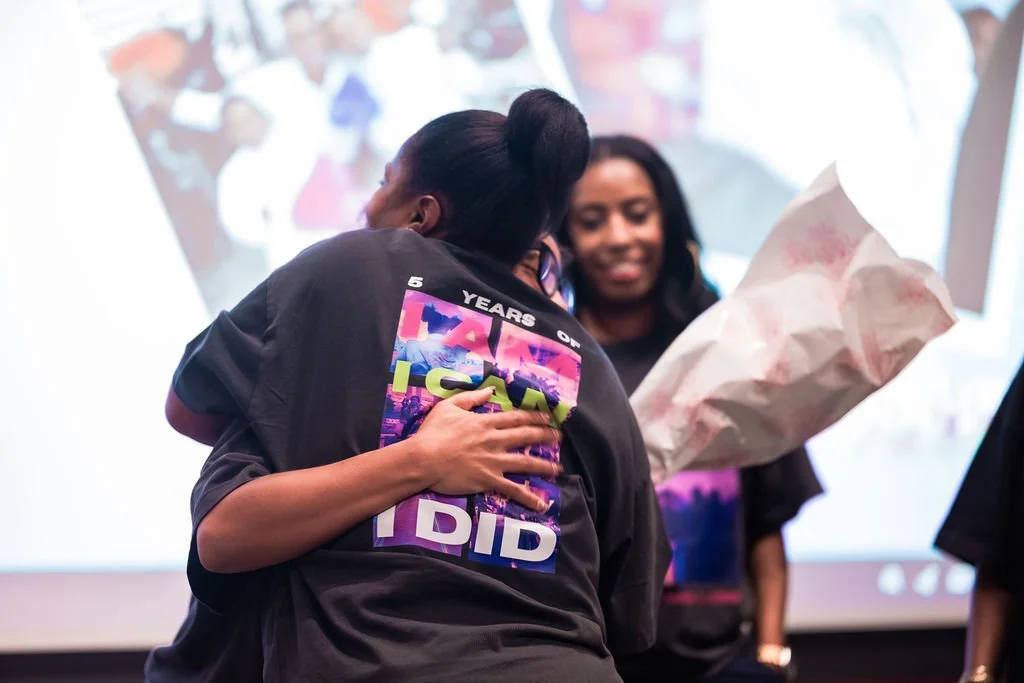 Two women hugging during a celebration, with one holding a large bouquet of flowers, at an event in front of a screen.
