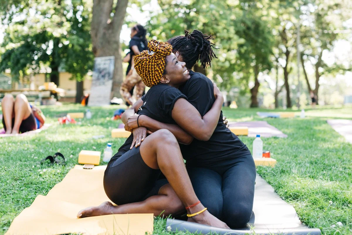 Two women hugging each other warmly while kneeling on yoga mats outdoors in a park with green trees and grass on a sunny day.