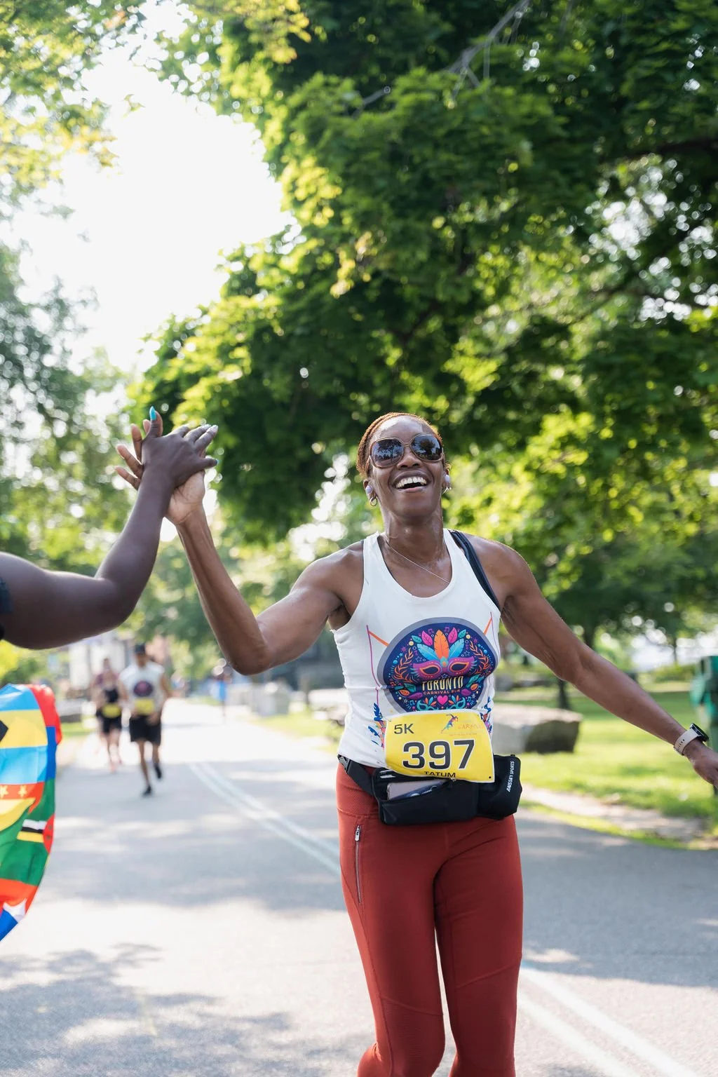 A woman wearing sunglasses and a tank top with a colorful skull logo, smiling and high-fiving someone during a 5K race on a sunny day with green trees in the background.