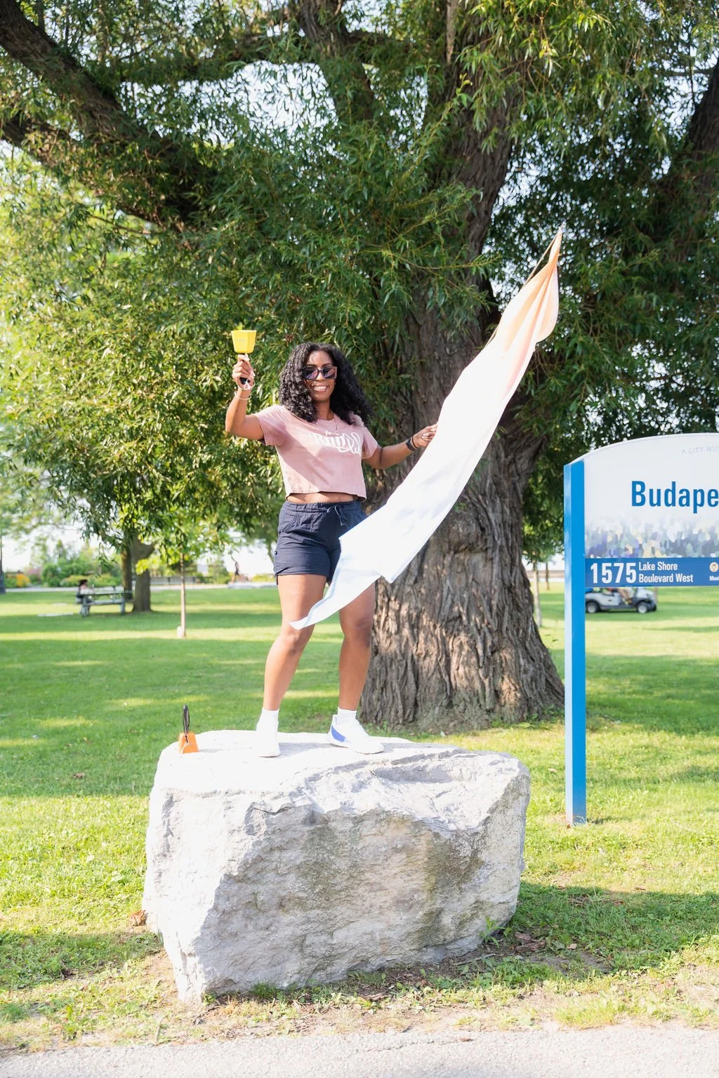 A young woman with sunglasses smiling and holding a small yellow trowel, standing on a large rock near a sign that reads "Budap" in a park. She is wearing a pink crop top, dark shorts, and white shoes. She is adjusting a white flag or banner next to 