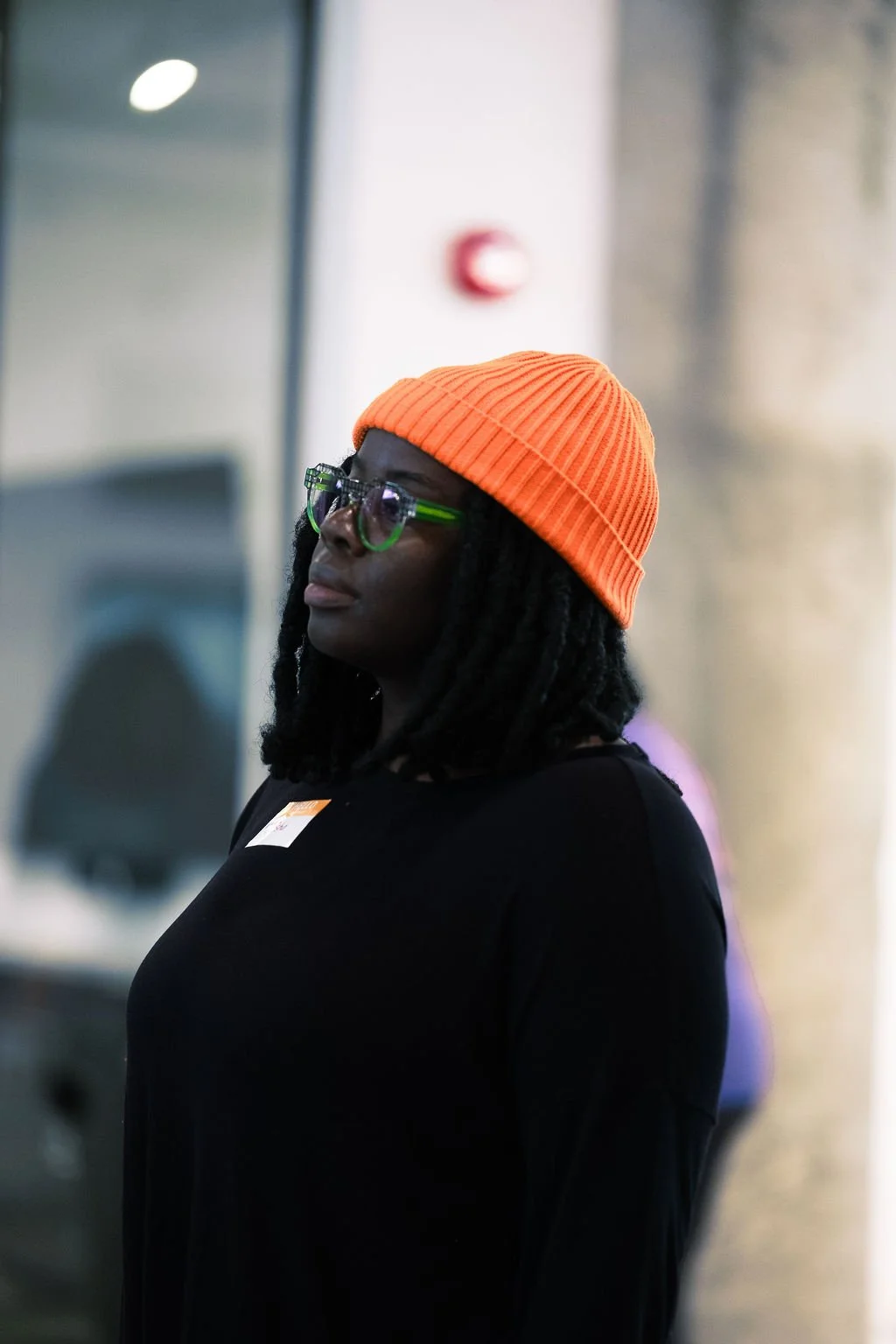 A woman with dark skin wearing an orange knit beanie and large green glasses, standing indoors with a focused expression.