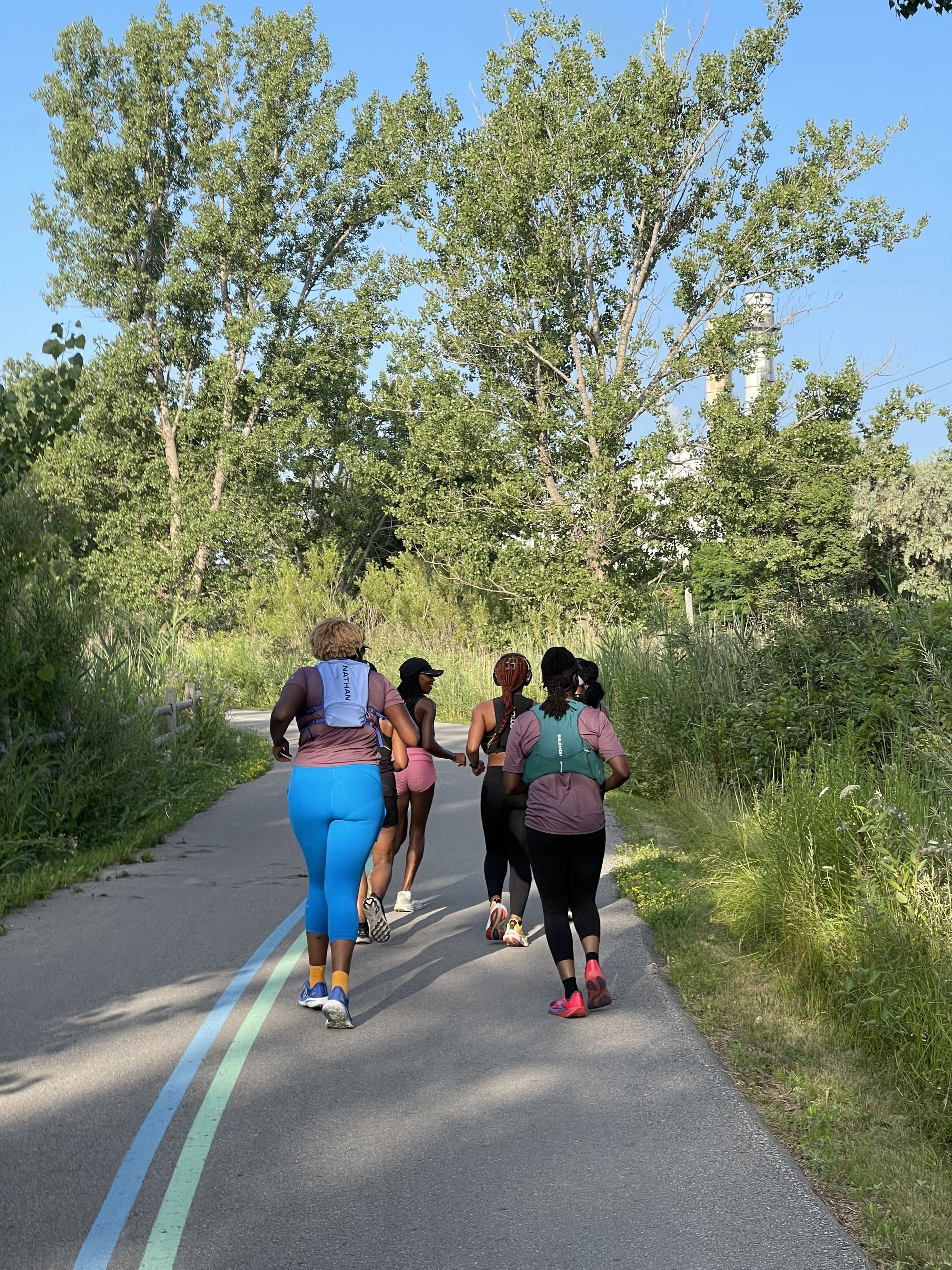 Group of five women jogging on a shaded, tree-lined road under clear blue sky.
