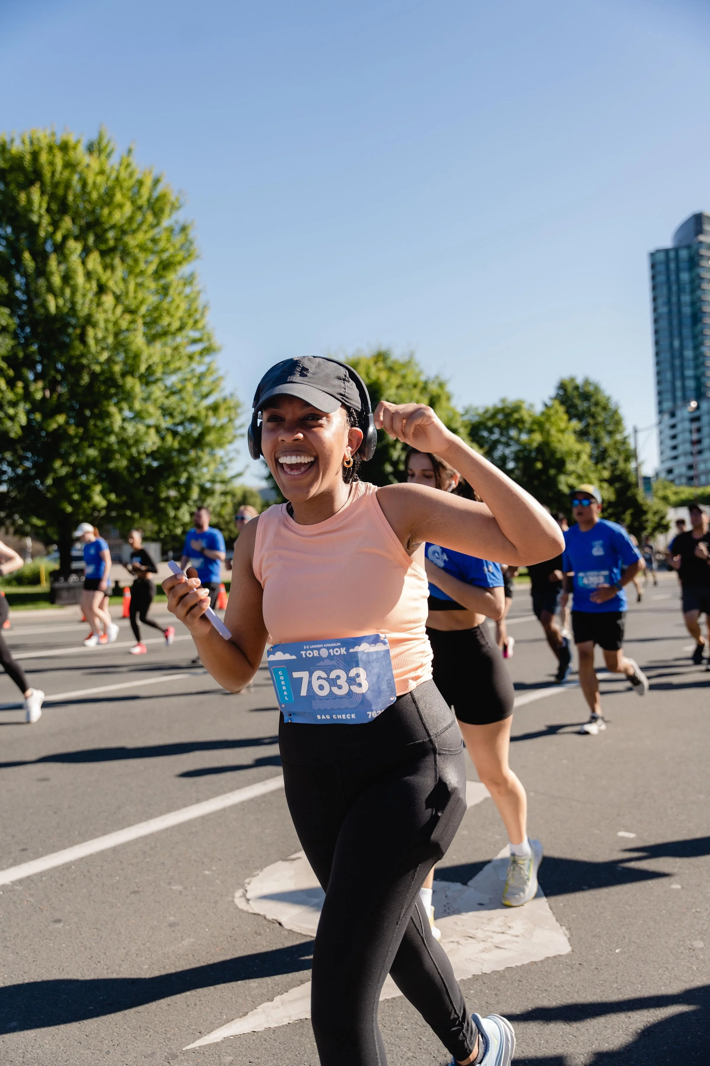 A woman wearing a black cap, headphones, and a peach tank top smiling while running in a marathon surrounded by other runners on a sunny city street with trees and tall buildings.