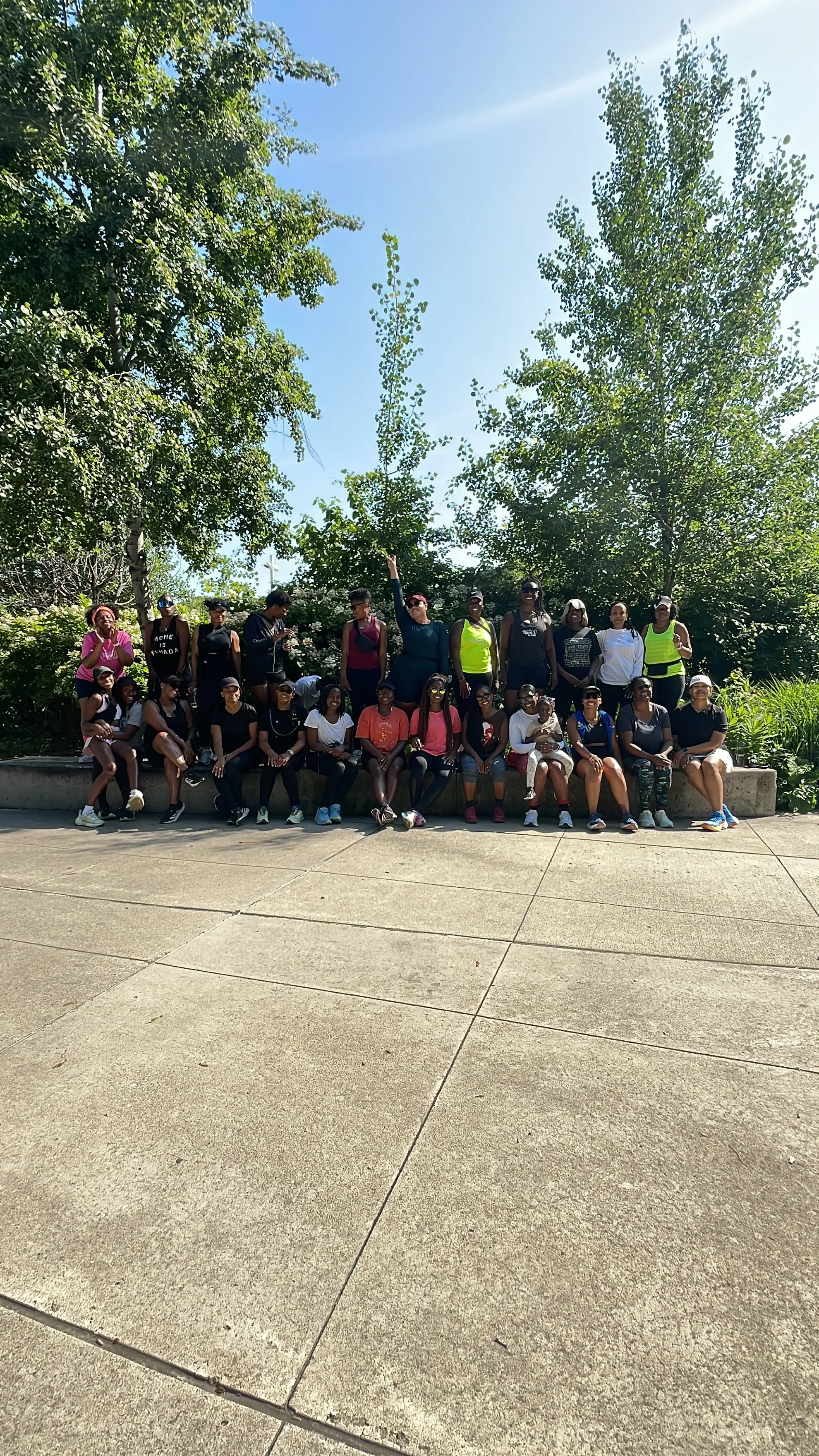 A large group of women and girls are gathered outdoors for a group photo, seated and standing on a concrete ledge with trees and greenery in the background on a sunny day.