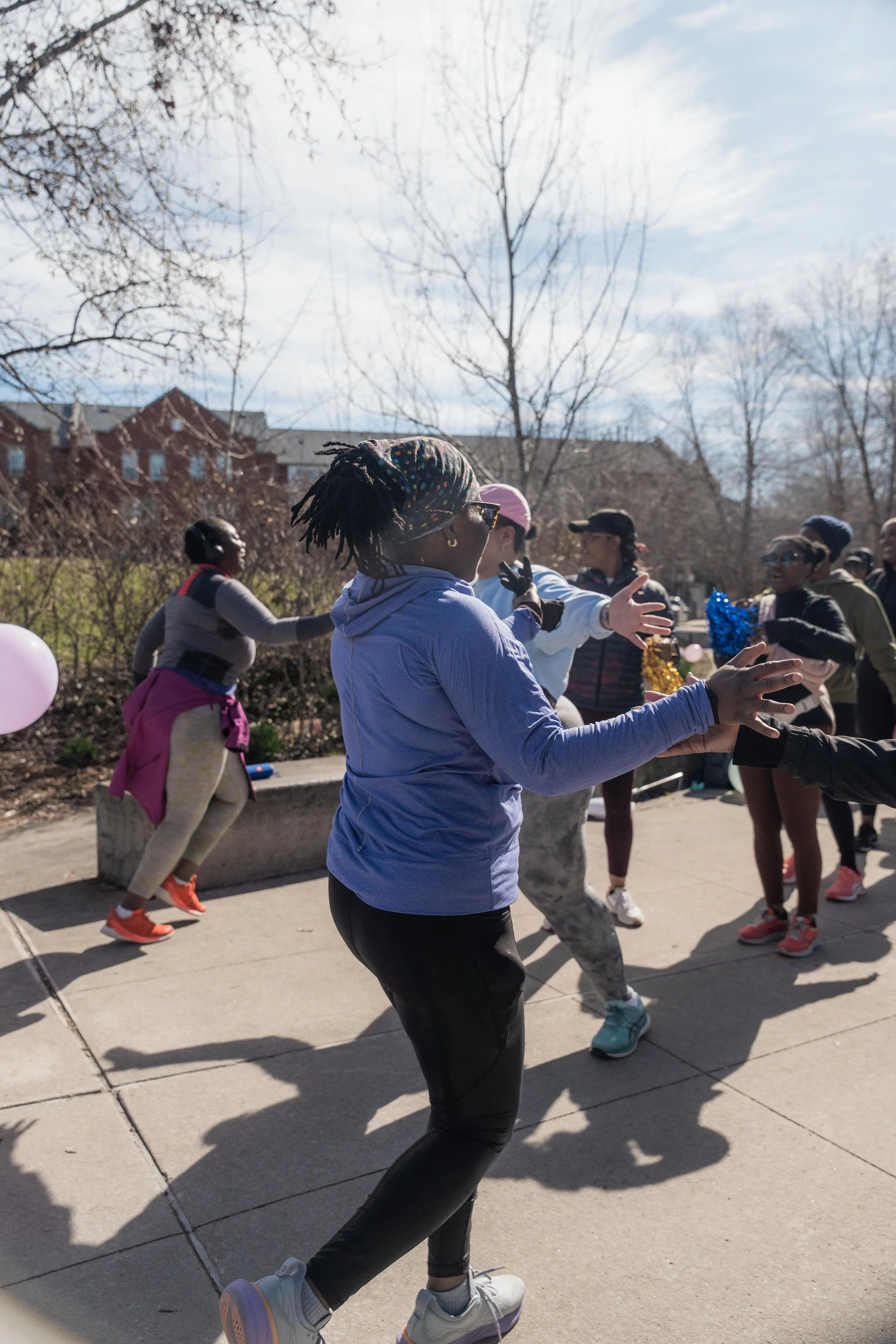 People participating in an outdoor activity, engaging in a group exercise or dance on a sidewalk with leafless trees and buildings in the background, under a partly cloudy sky.