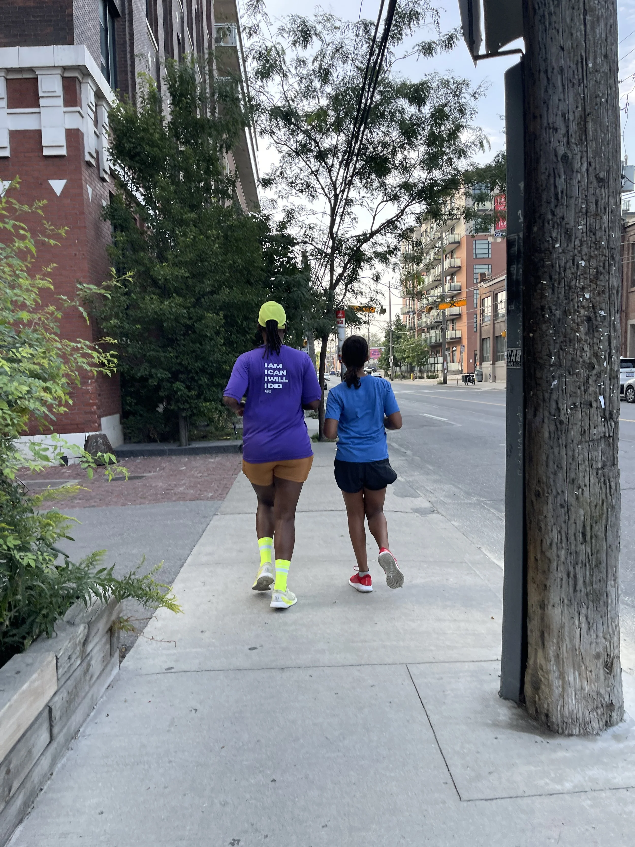 Two women are jogging on a sidewalk in an urban area with trees and multi-story buildings around. One woman is wearing a purple shirt and yellow shorts, and the other is wearing a blue shirt and black shorts.