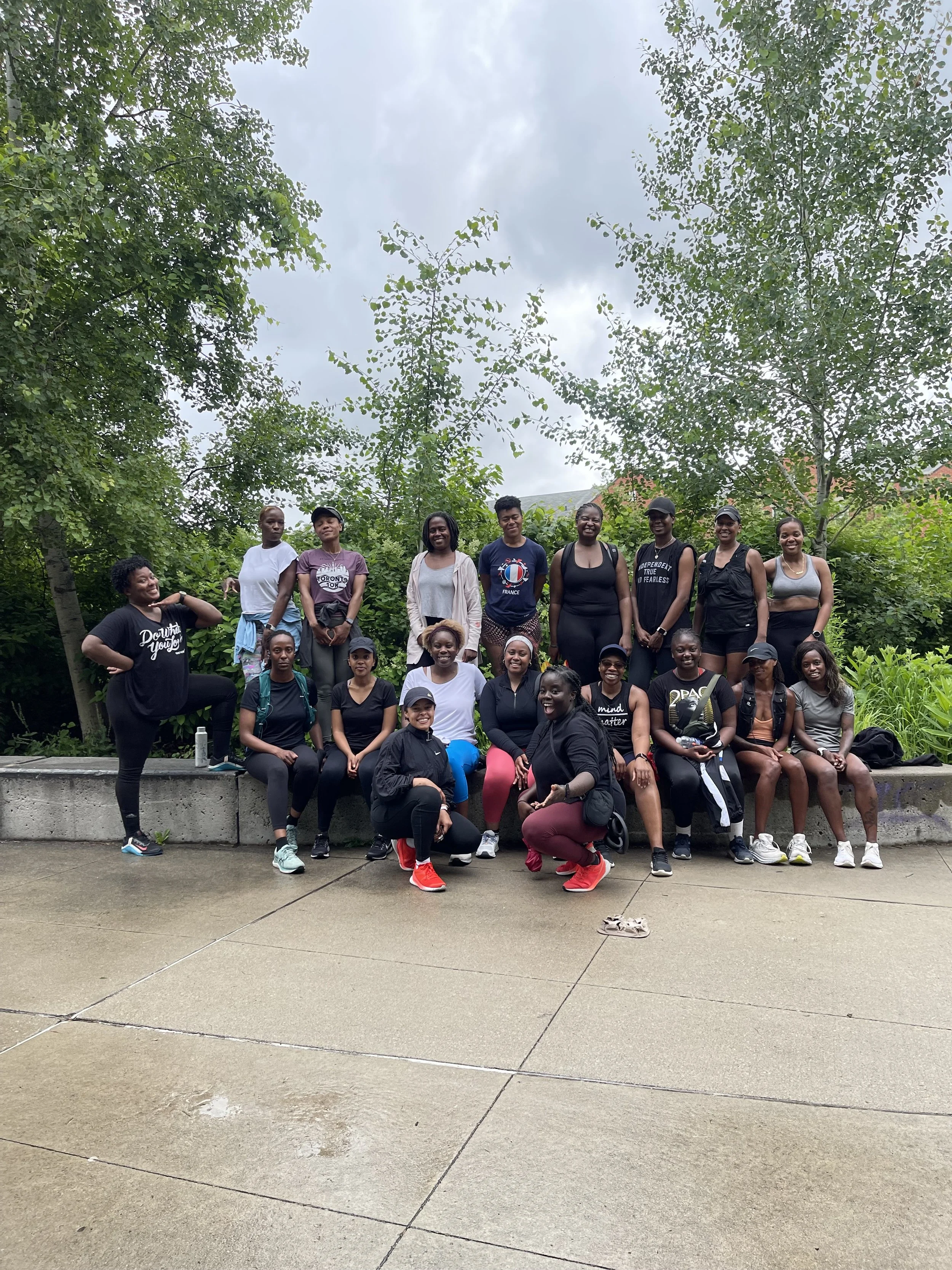 Group of people posing outdoors on a cloudy day, standing and sitting on a low concrete wall surrounded by trees and greenery.