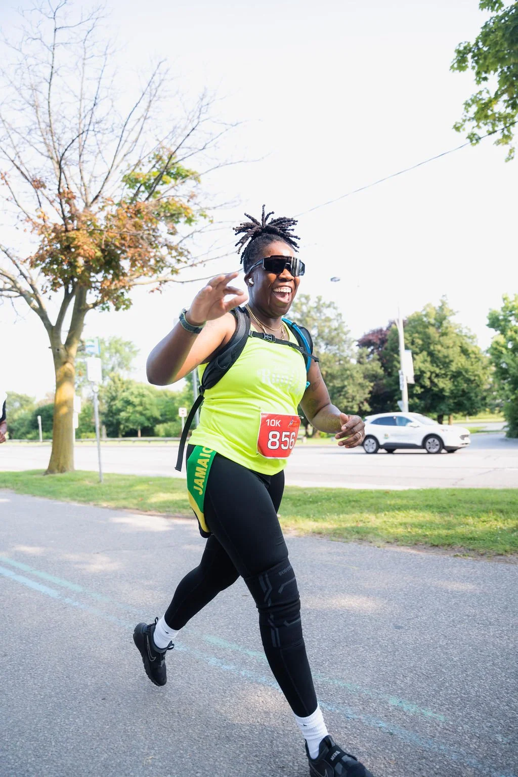 A woman running outdoors during a race, wearing a bright yellow race shirt with a bib numbered 856, black leggings, sunglasses, and a backpack, with trees and a car in the background.