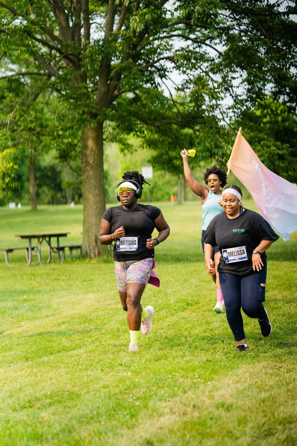 Three women run in a park during a race, with trees and a picnic table in the background. They are wearing athletic clothing, race bibs, and accessories, with one woman holding a pink flag.