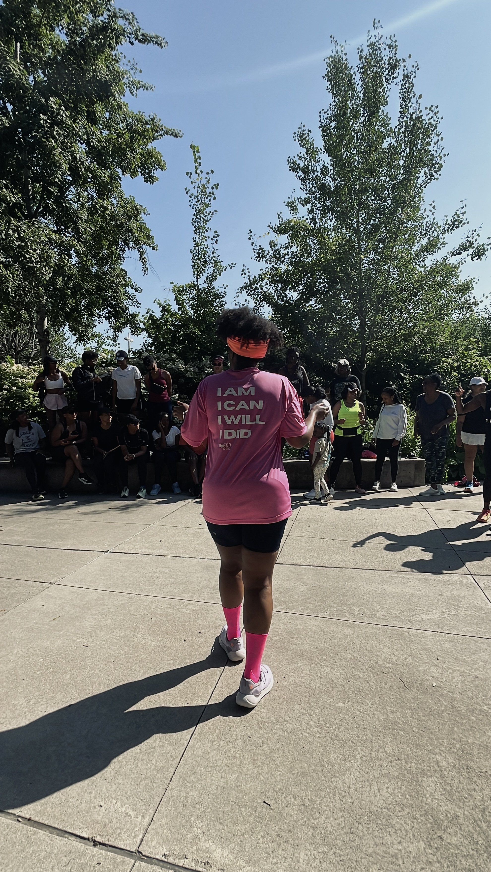 Person with curly hair wearing a pink shirt with motivational text on the back, black shorts, pink socks, and white sneakers, standing outdoors on a concrete pavement, with a group of people sitting and standing in the background and trees under a cl