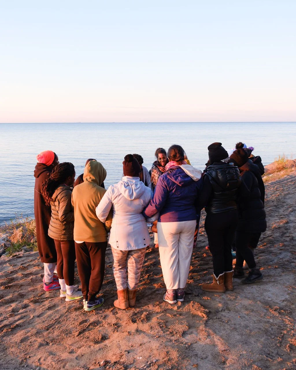 Group of diverse people huddled together on a sandy beach at sunset, near a calm body of water, sharing a moment of togetherness.