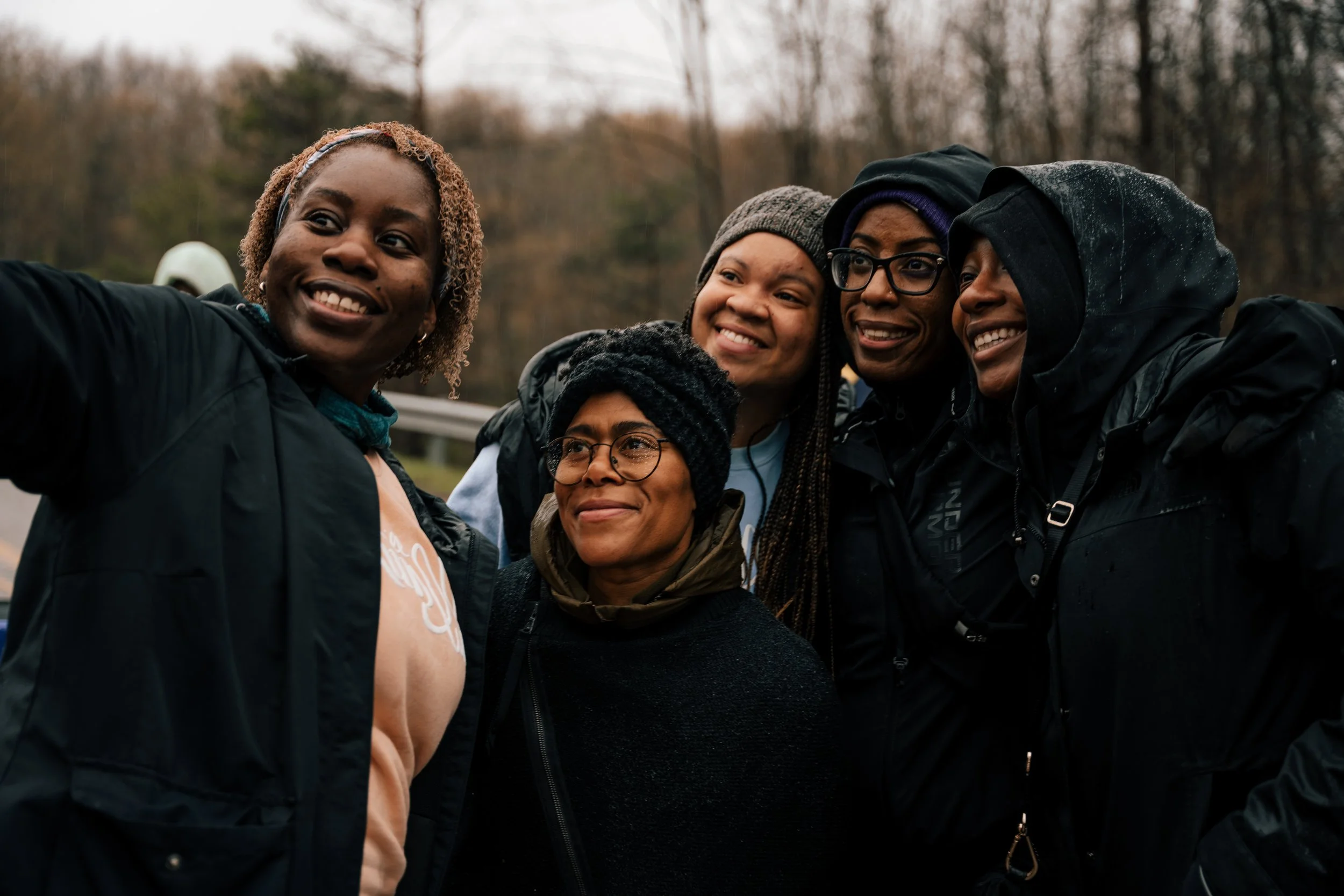 Five women outdoors taking a selfie together in a wooded area during overcast weather.