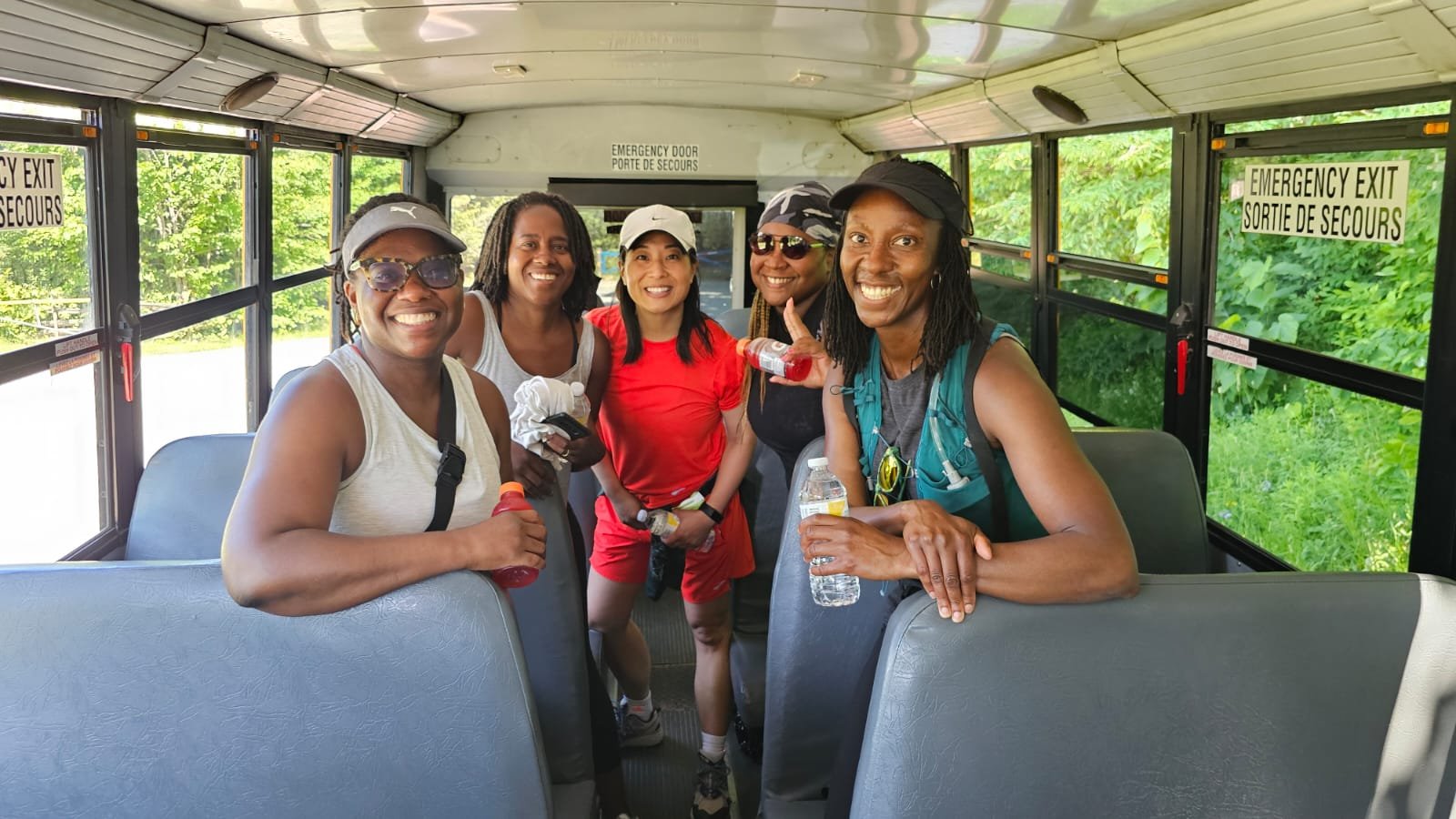 Group of five women on a bus, smiling and holding water bottles, dressed in casual outdoor athletic clothing, with trees visible outside the bus windows.