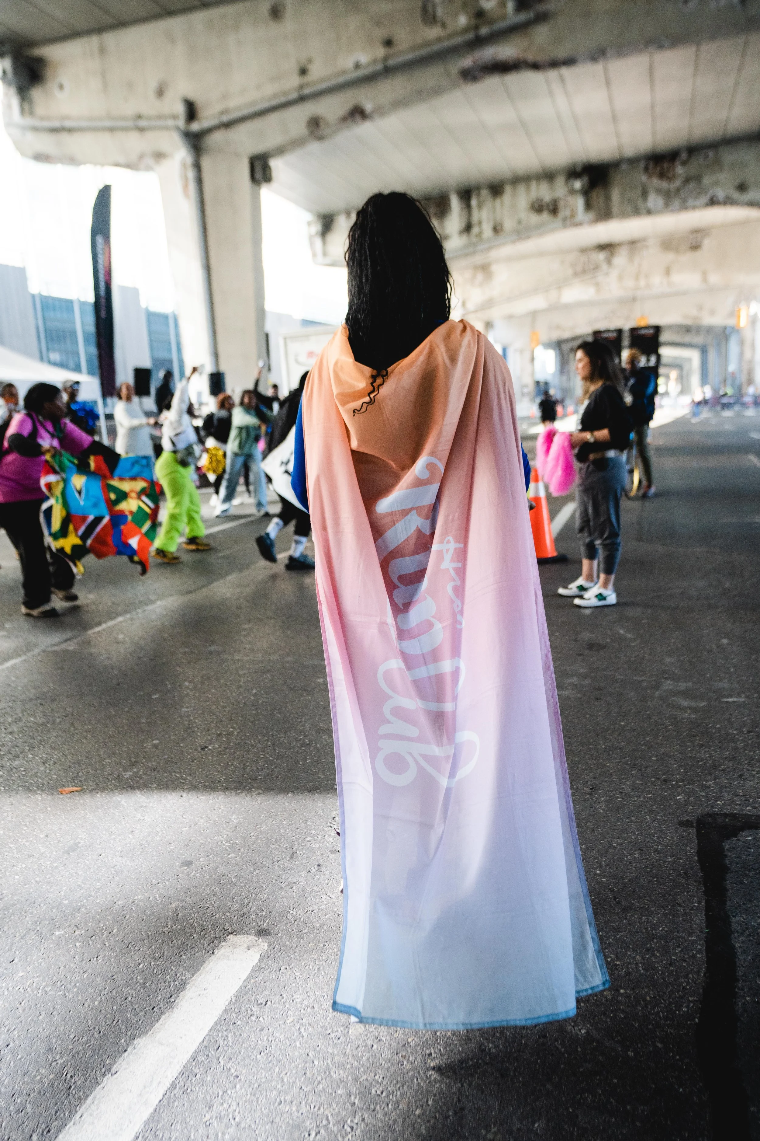 A person wearing a rainbow-colored pride flag as a cape at an outdoor event under a bridge, with a crowd of people around them.