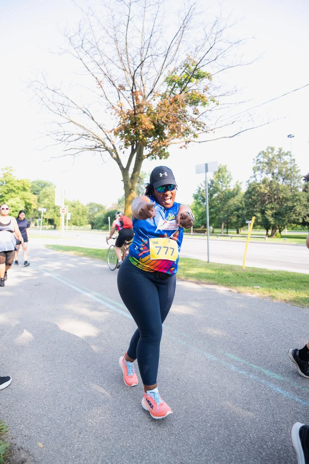 Woman in athletic wear and sunglasses celebrating during a 5K race, pointing towards the camera with a big smile, with other runners and a park setting in the background.