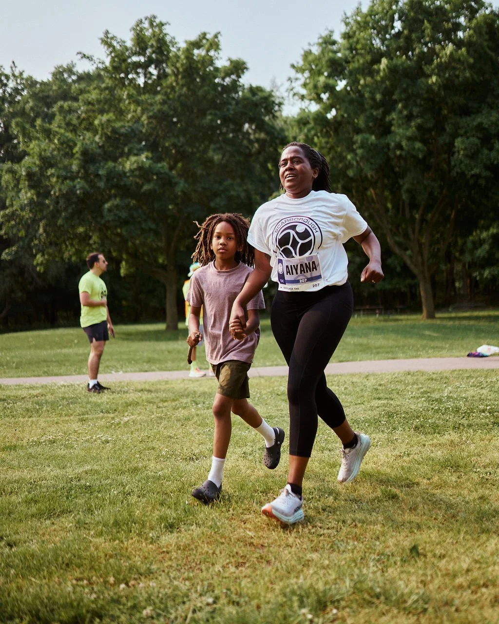 A woman and a young girl with dreadlocks holding hands and running in a park, participating in a race, with others in the background and trees surrounding the area.