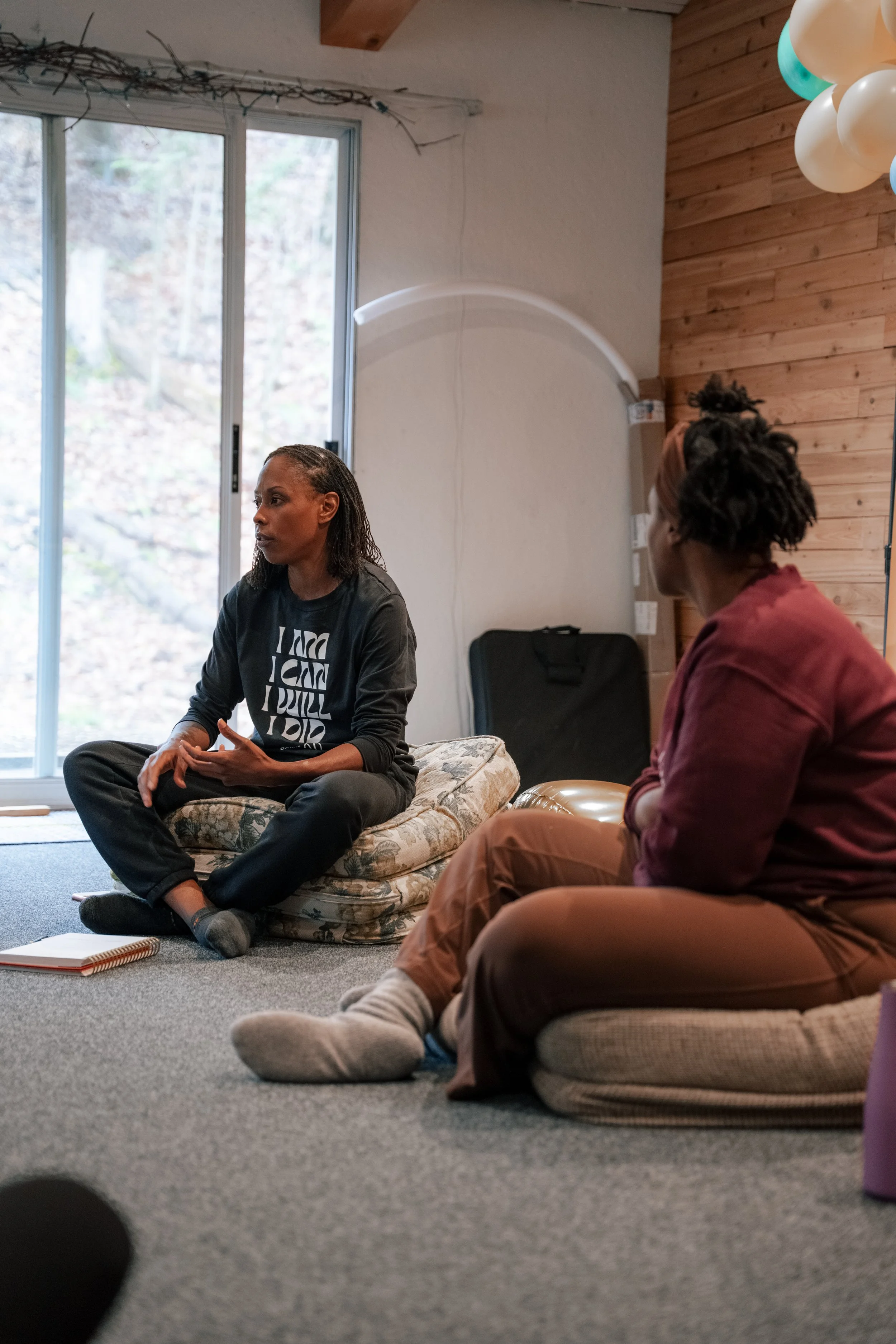 Two women sitting on cushions on the floor of a room with large windows and wooden wall paneling, engaging in a conversation.