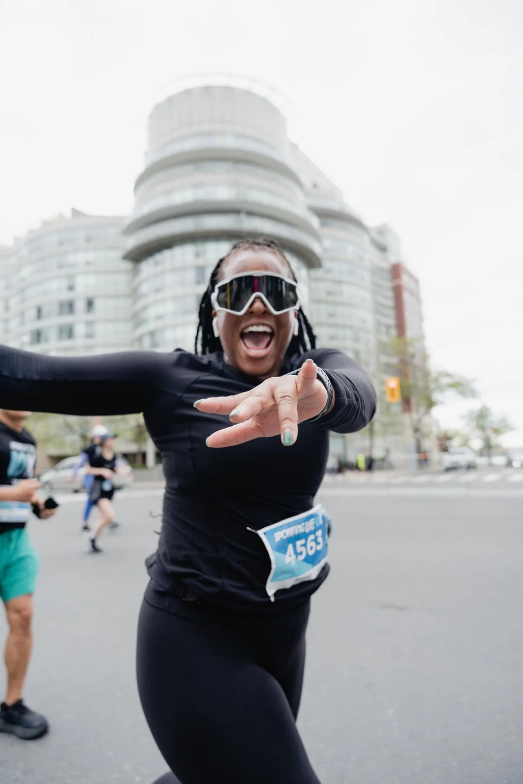 Female marathon runner smiling and reaching out during a race, wearing sunglasses and a black athletic outfit, with a city building in the background.