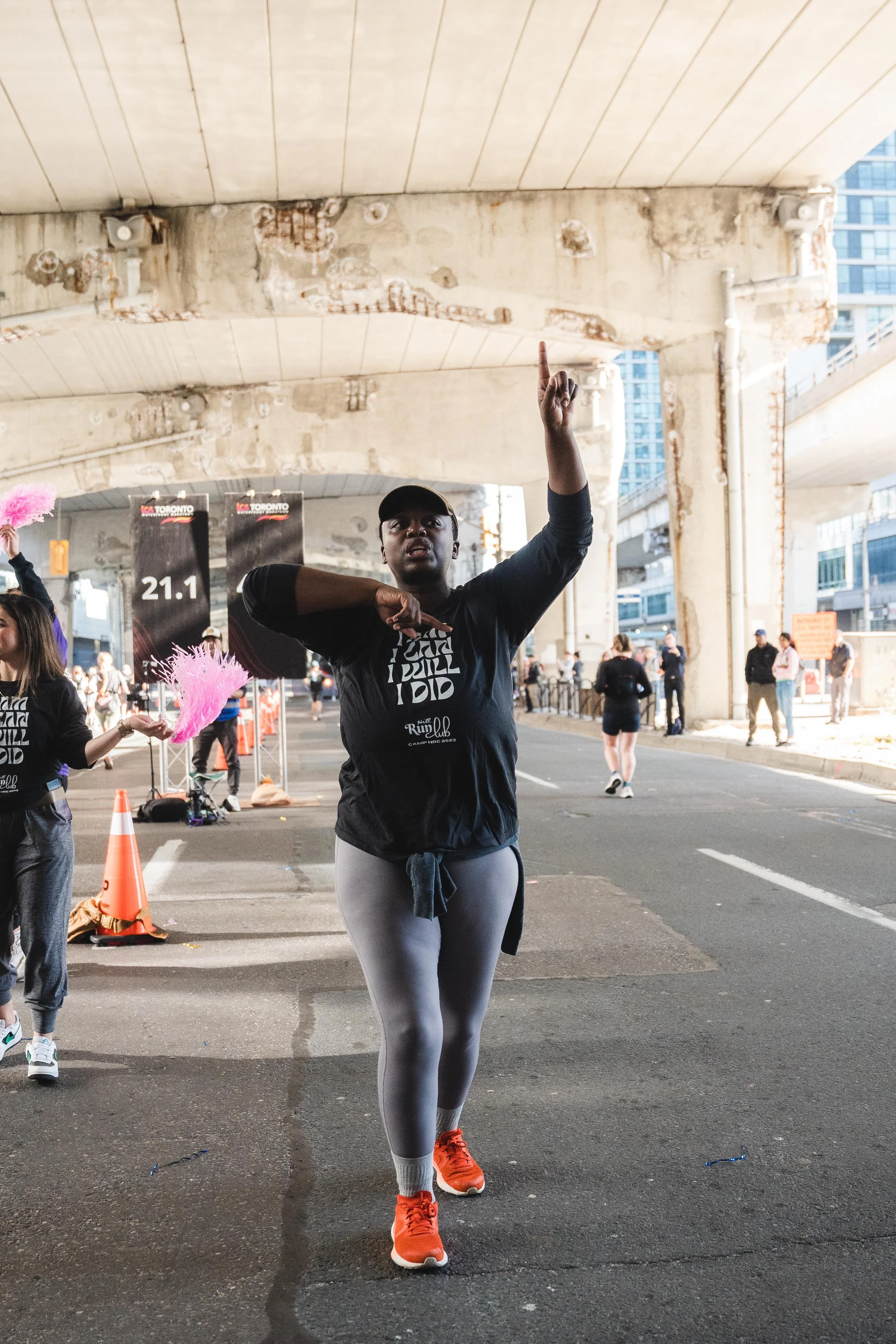 A woman in athletic clothing is dancing or performing in the middle of a street during a marathon event under an overpass, with other participants and spectators in the background.