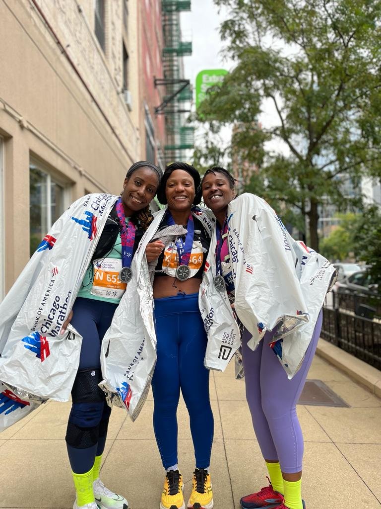 Three Black women from hill run club, after completing the Chicago Marathon with medals, wrapped in race blankets, celebrate after a marathon in Chicago in an urban area with buildings and trees.
