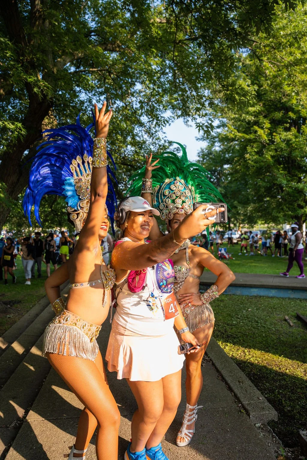 Three women in carnival costumes taking a selfie in a park with trees in the background. Two of the women are dressed in colorful feathered headdresses and costumes adorned with jewels, while the third woman in the center is in casual clothing.