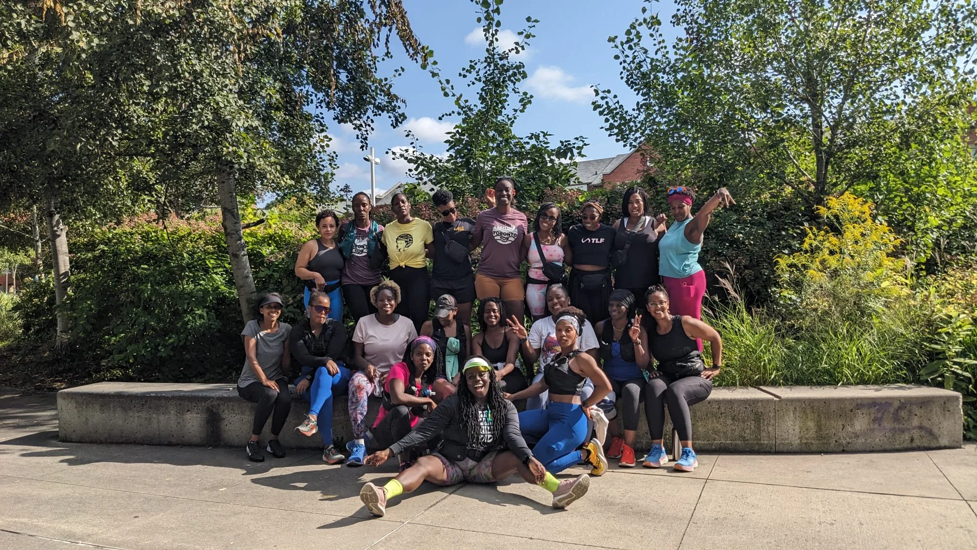 Group of women outdoors on a sunny day, posing together in front of greenery. Some are seated, others standing, with a clear blue sky overhead.