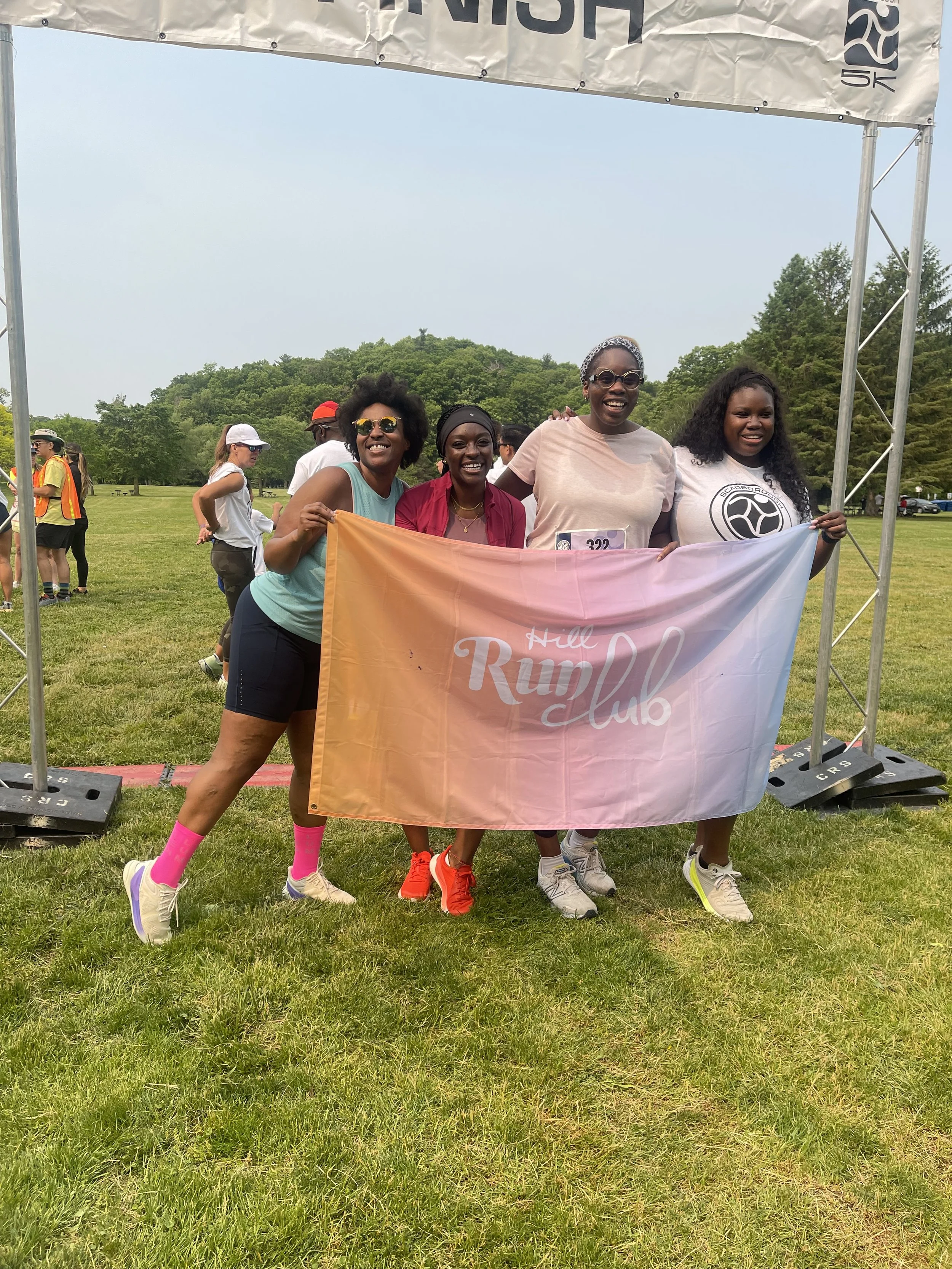 Four women standing on grass holding a pink and orange banner that reads 'Hill Rump Club' at an outdoor event, smiling and posing for the picture.