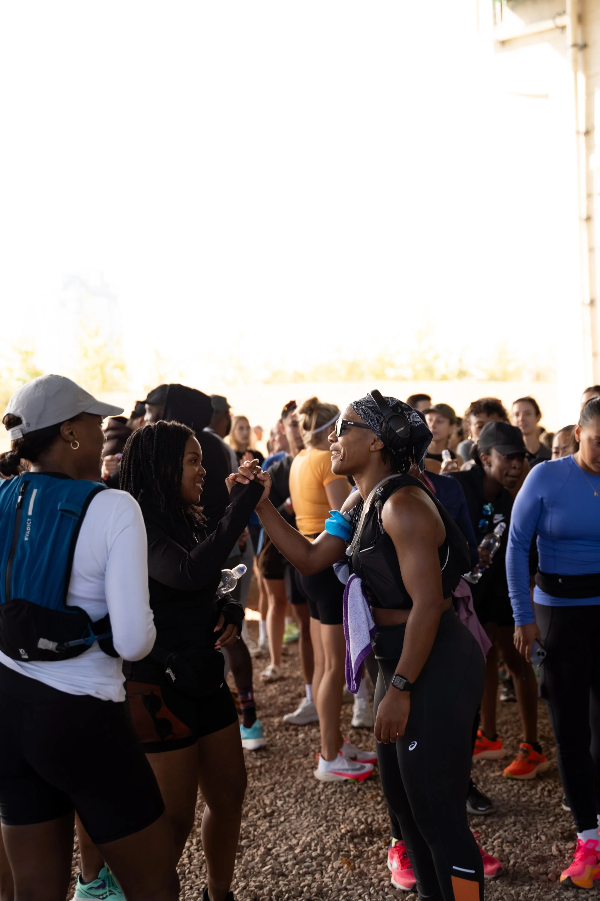 Group of women in athletic clothing gathered outdoors, greeting each other cheerfully, some holding water bottles.