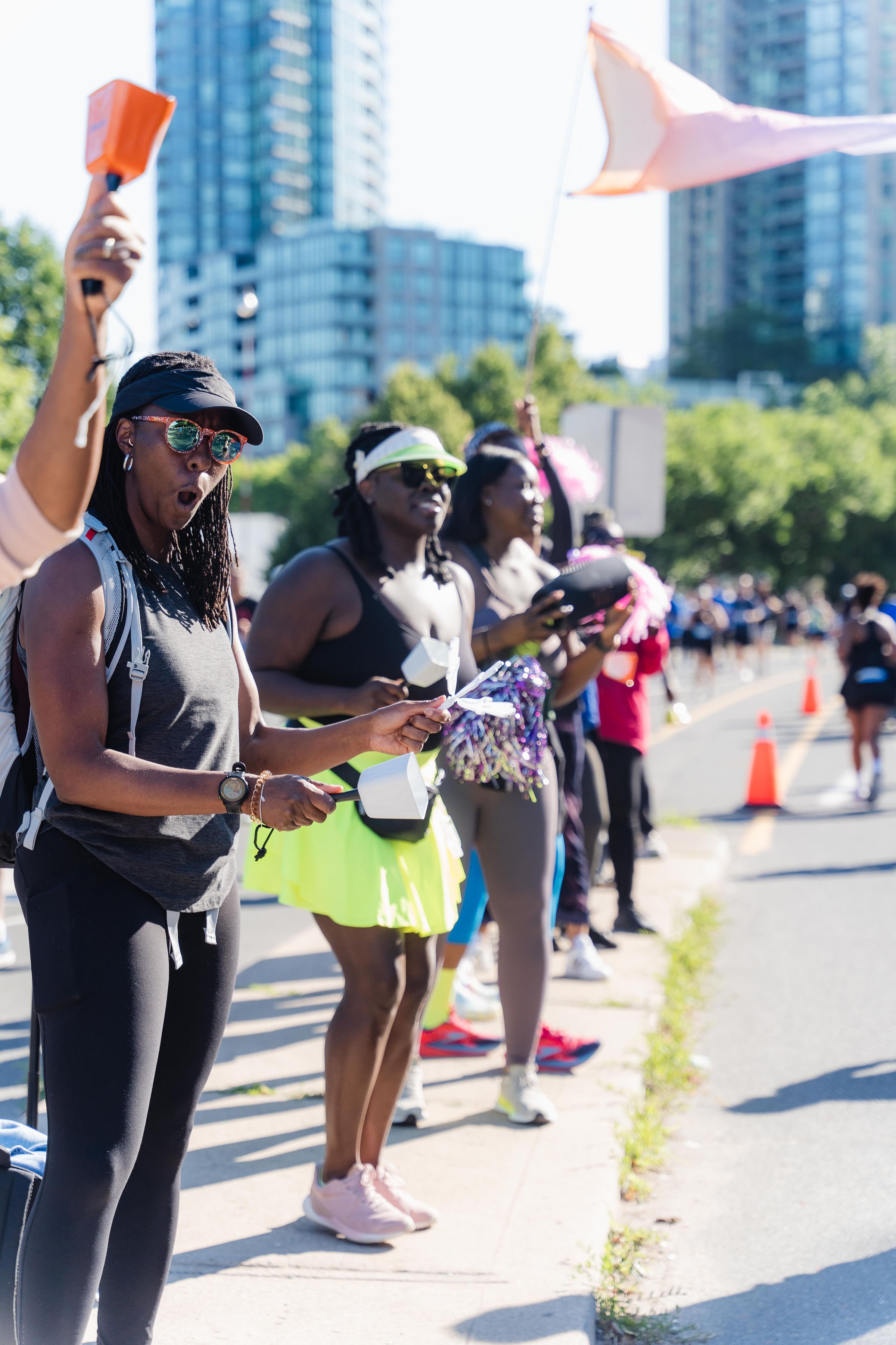 Women standing on the side of a street during a race or marathon, holding whistles and cheering, with orange cones and high-rise buildings in the background on a sunny day.