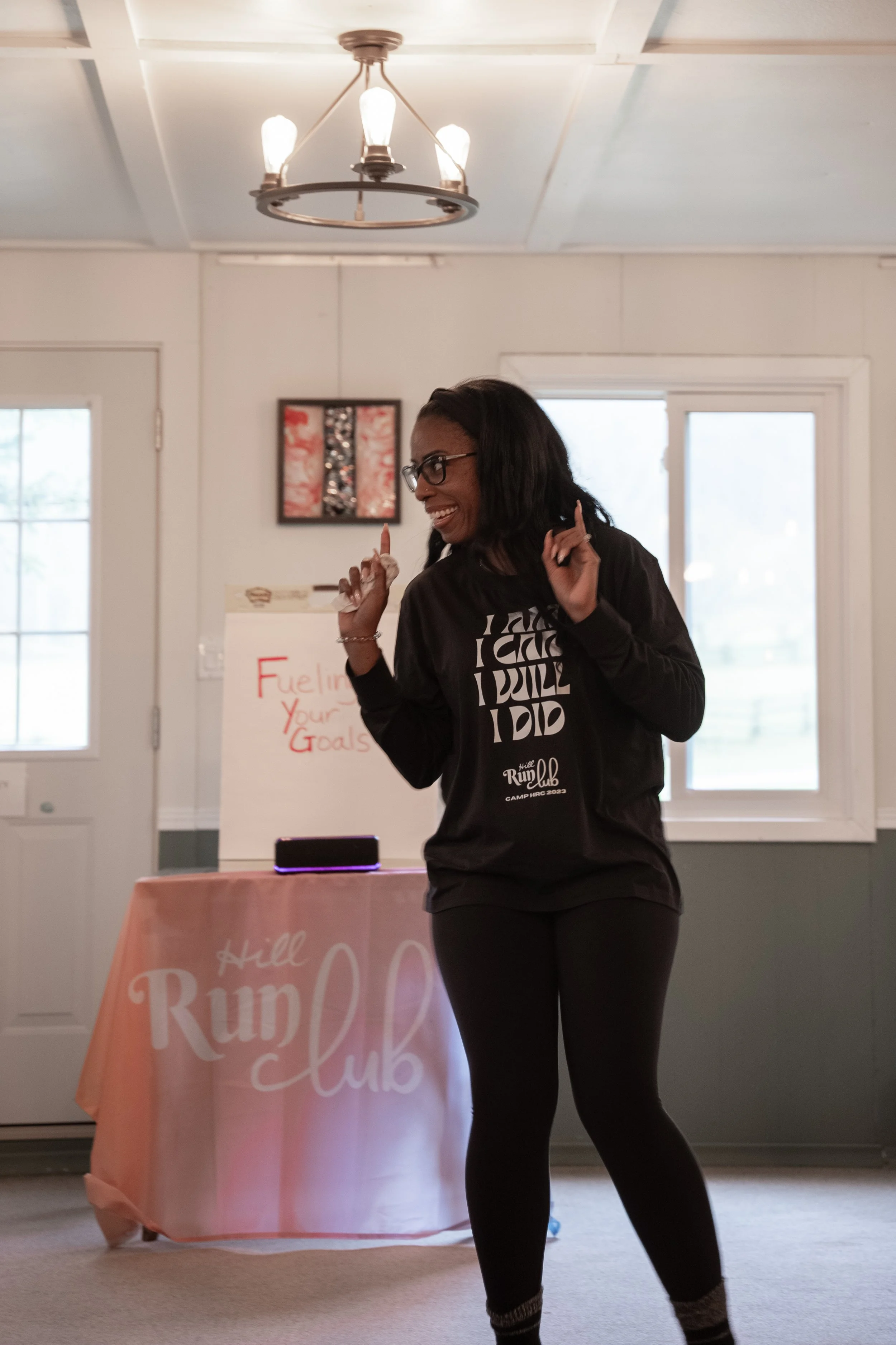 A woman with dark hair and glasses dancing indoors at Hill Run Club event, wearing a black sweatshirt and black leggings.