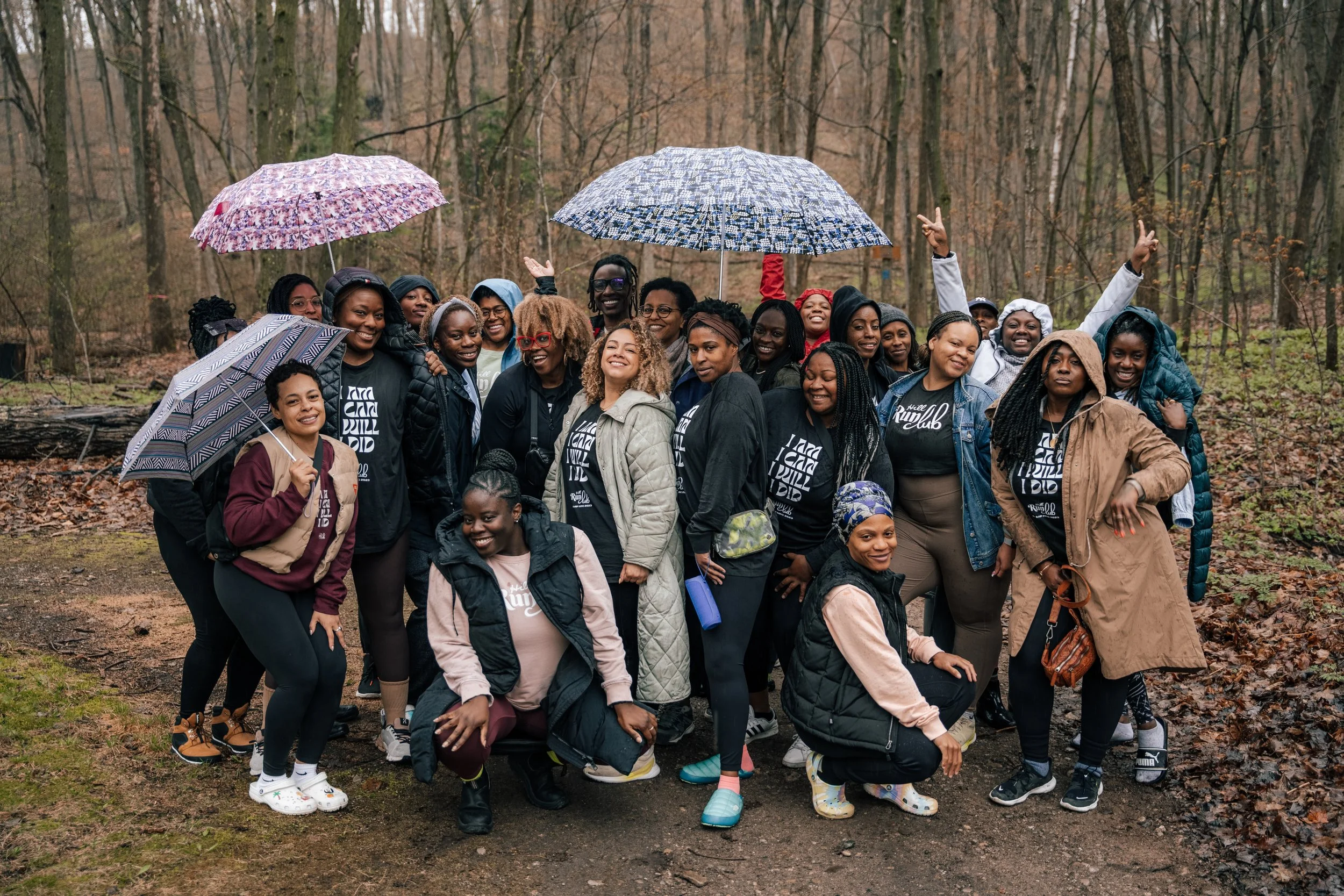 Group of women smiling and posing together in a wooded outdoor area during rainy weather, some holding umbrellas.