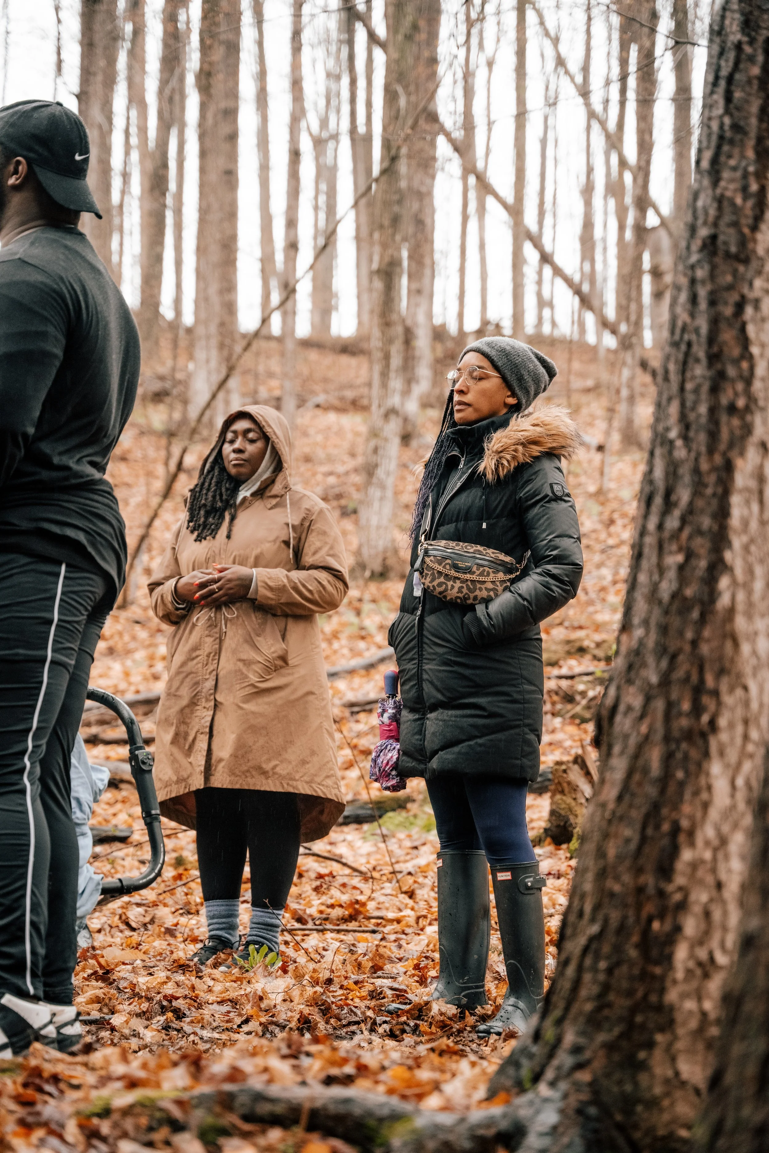 Three women and a man standing in a forest surrounded by fallen autumn leaves, with trees in the background; the women are wearing winter coats and rubber boots, and appear to be deep in thought or prayer.